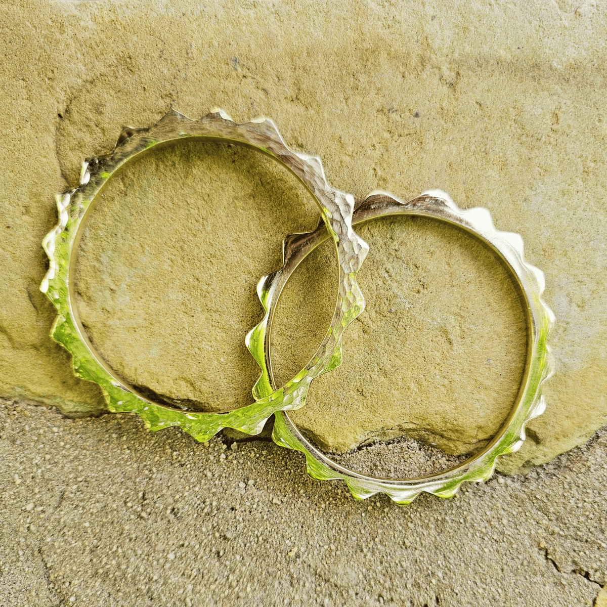 Two gold-toned metal bangle bracelet with a textured, spiked outer edge, resting on a natural stone surface with a blurred outdoor background. Shop Kamawe