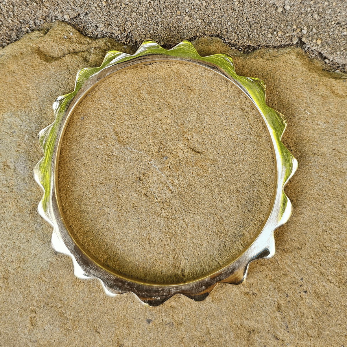 A circular metal ring with a scalloped, wavy edge, filled with a fine tan powder, photographed from above on a sandy surface. Shop Kamawe