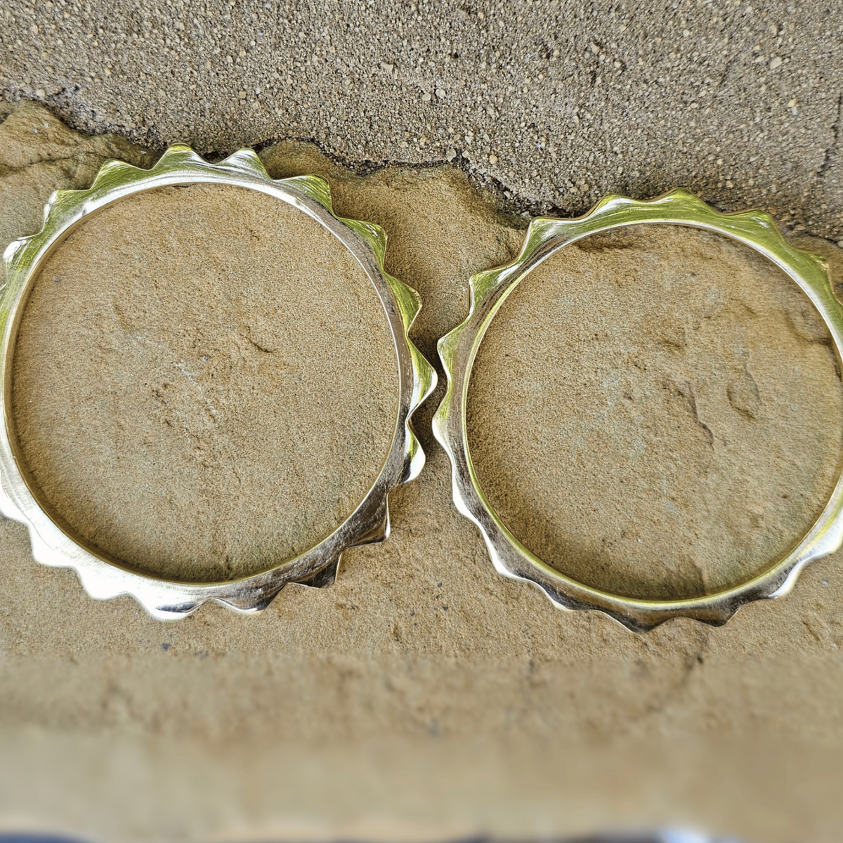 Two silver metal bottle cap rings placed side by side on a textured sandy beige surface. Shop Kamawe