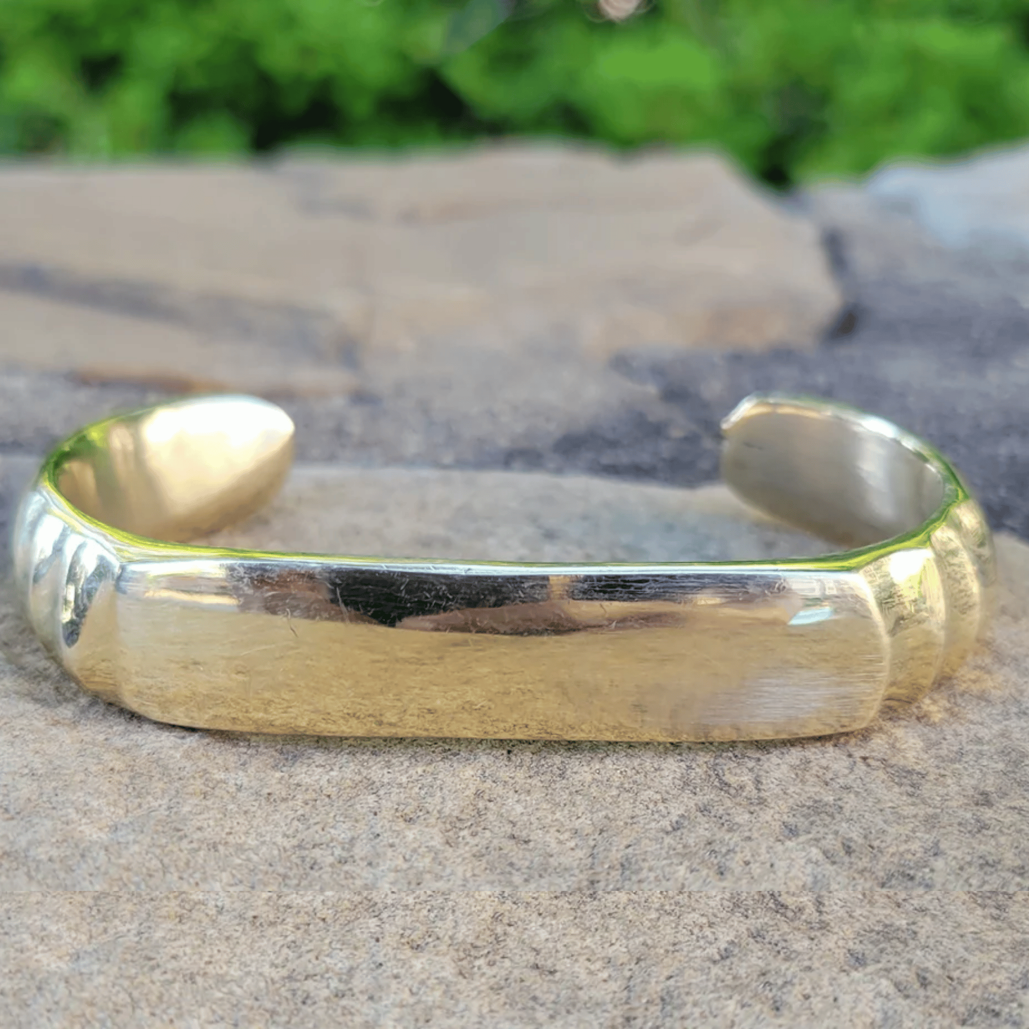 A polished gold cuff bracelet resting on a textured stone surface with a blurred green outdoor background. Shop Kamawe