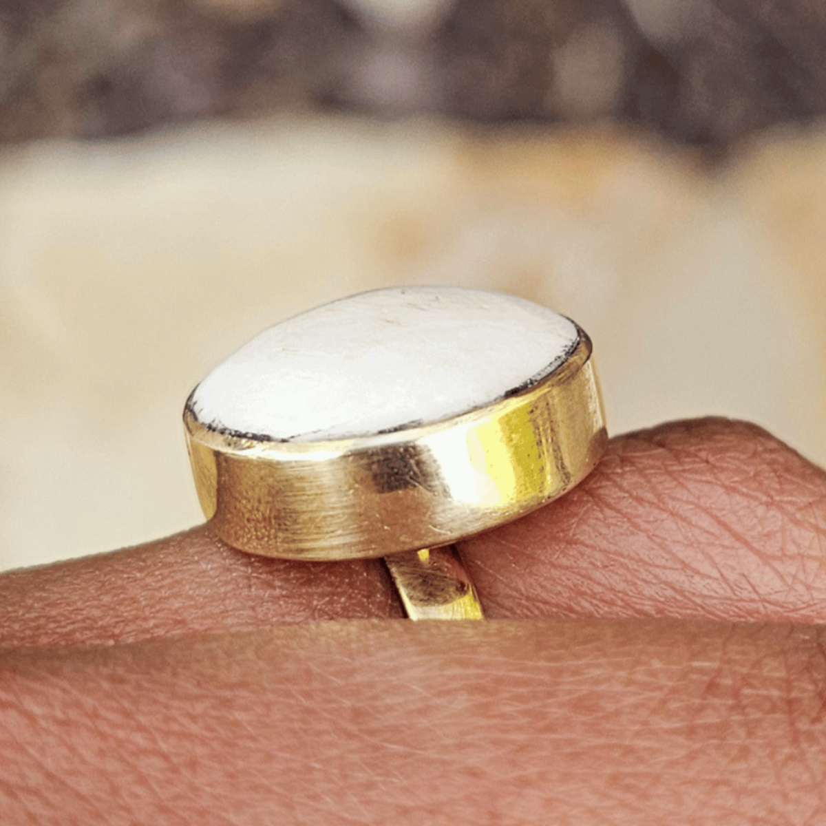 A gold ring with a large, oval white stone worn on a person's finger, set against a blurred background of earthy tones. Shop Kamawe