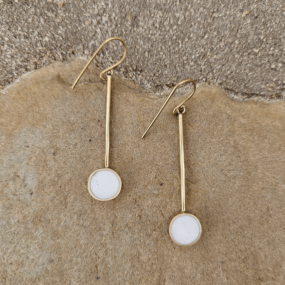 A pair of gold drop earrings with round white inlay discs, displayed on a textured beige stone surface. Shop Kamawe