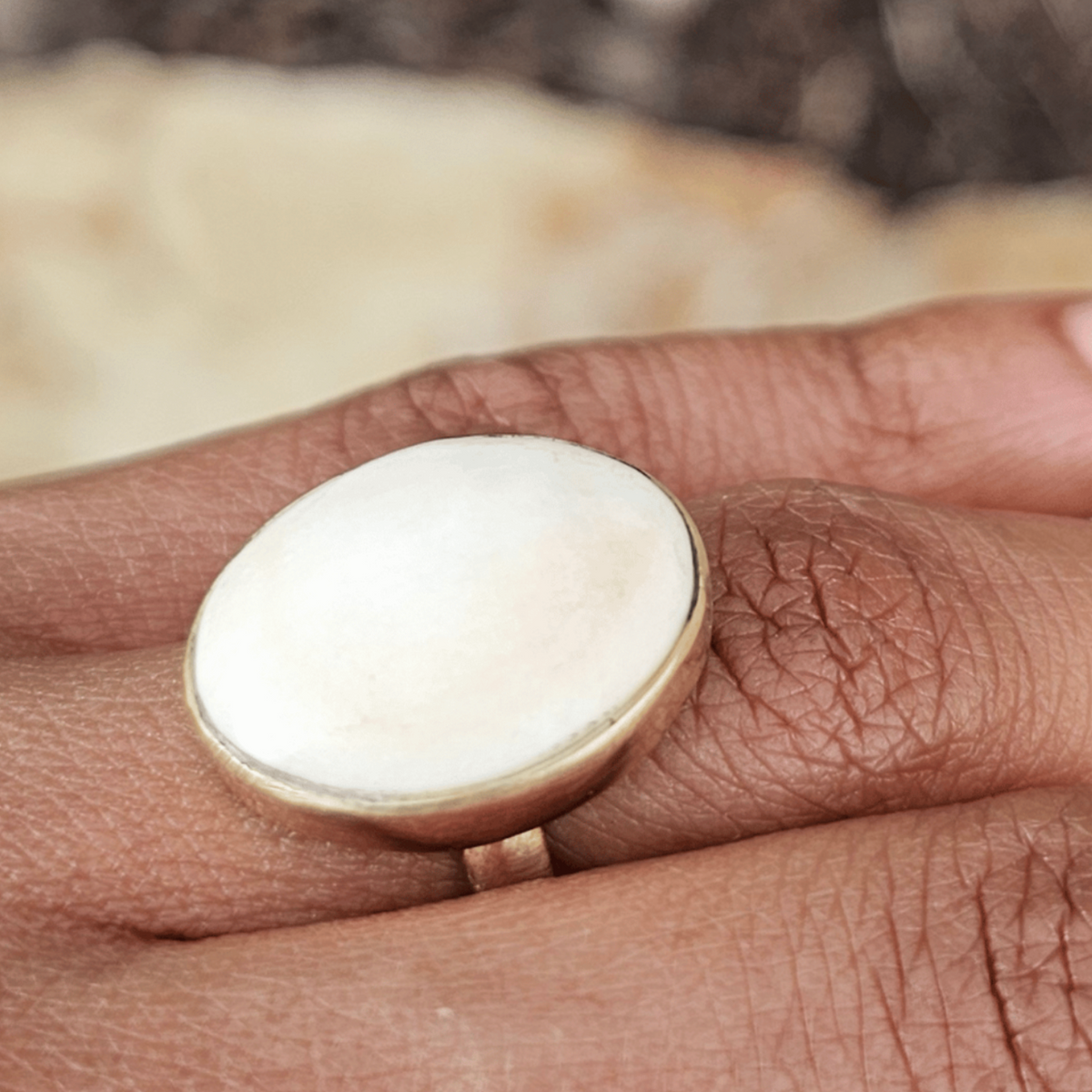 A close-up of a hand wearing a large oval ring with a cream-colored stone and a gold-toned setting, against a blurred natural background. Shop Kamawe