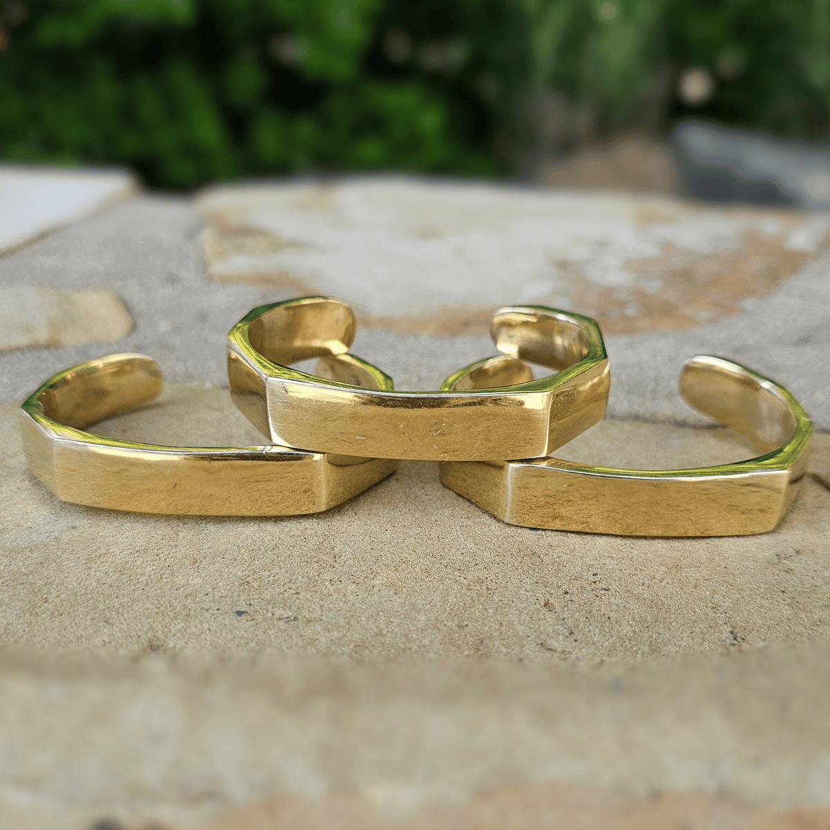 Three gold-toned open cuff bracelets with a textured, geometric hexagonal design, arranged on a light stone surface with a blurred green background. Shop Kamawe