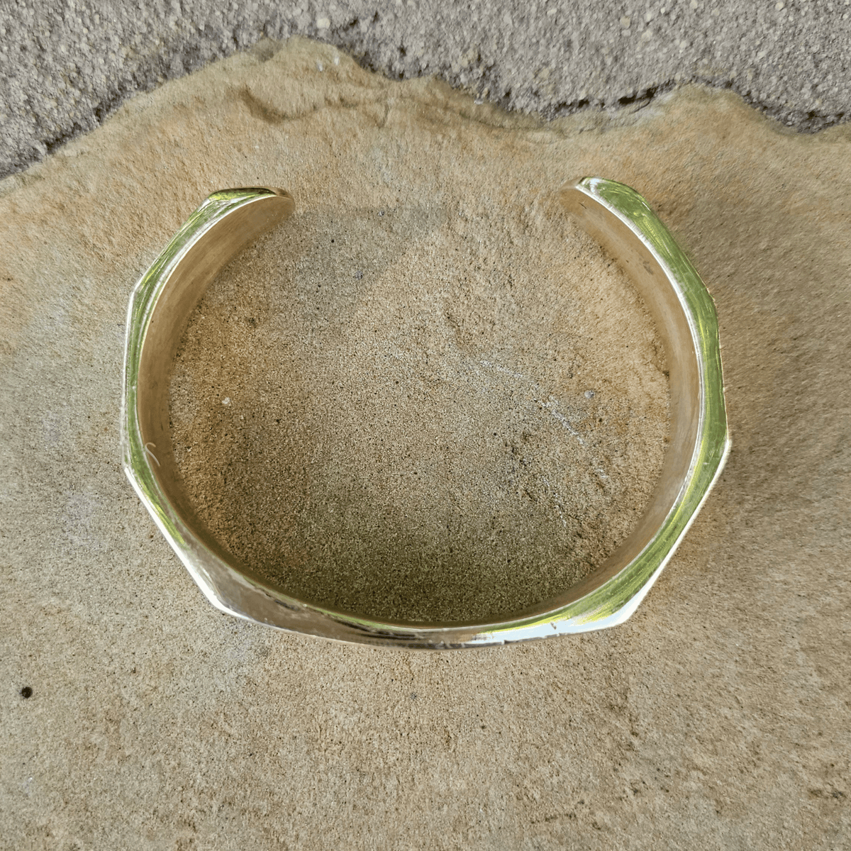 A chunky, open cuff bracelet with a polished gold finish, displayed on a textured beige surface. Shop Kamawe