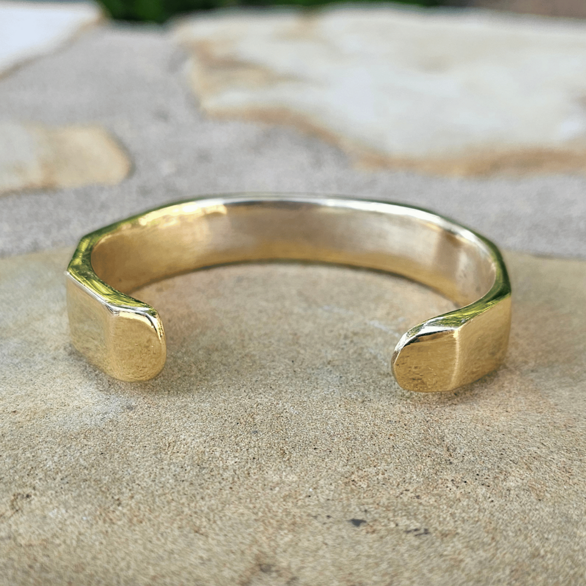 A gold-toned open cuff bracelet with a chunky, geometric, faceted design, resting on a textured stone surface outdoors. Shop Kamawe