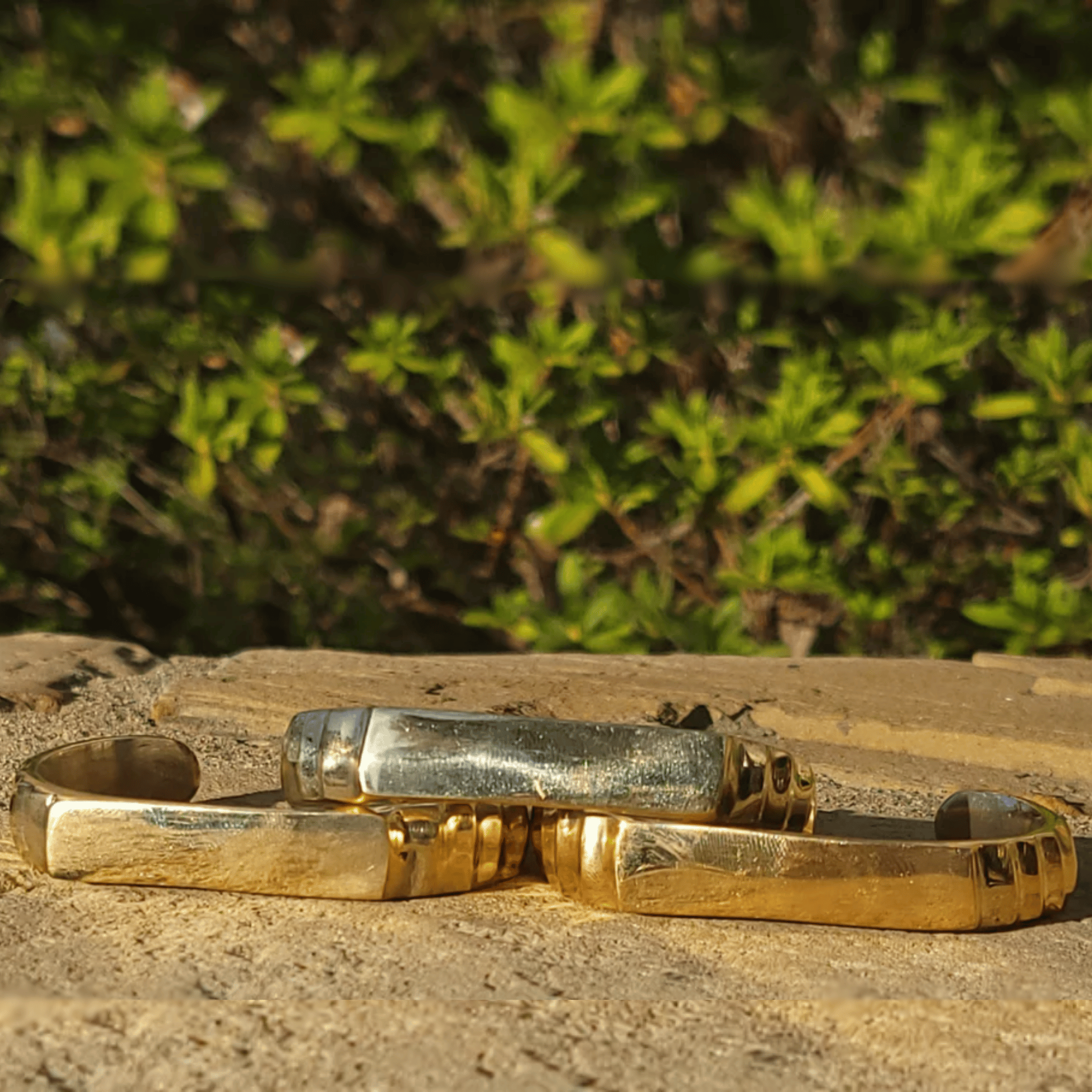Two metallic cuff bracelets, one gold-toned and one silver-toned, resting on a stone surface with a blurred green foliage background. Shop Kamawe