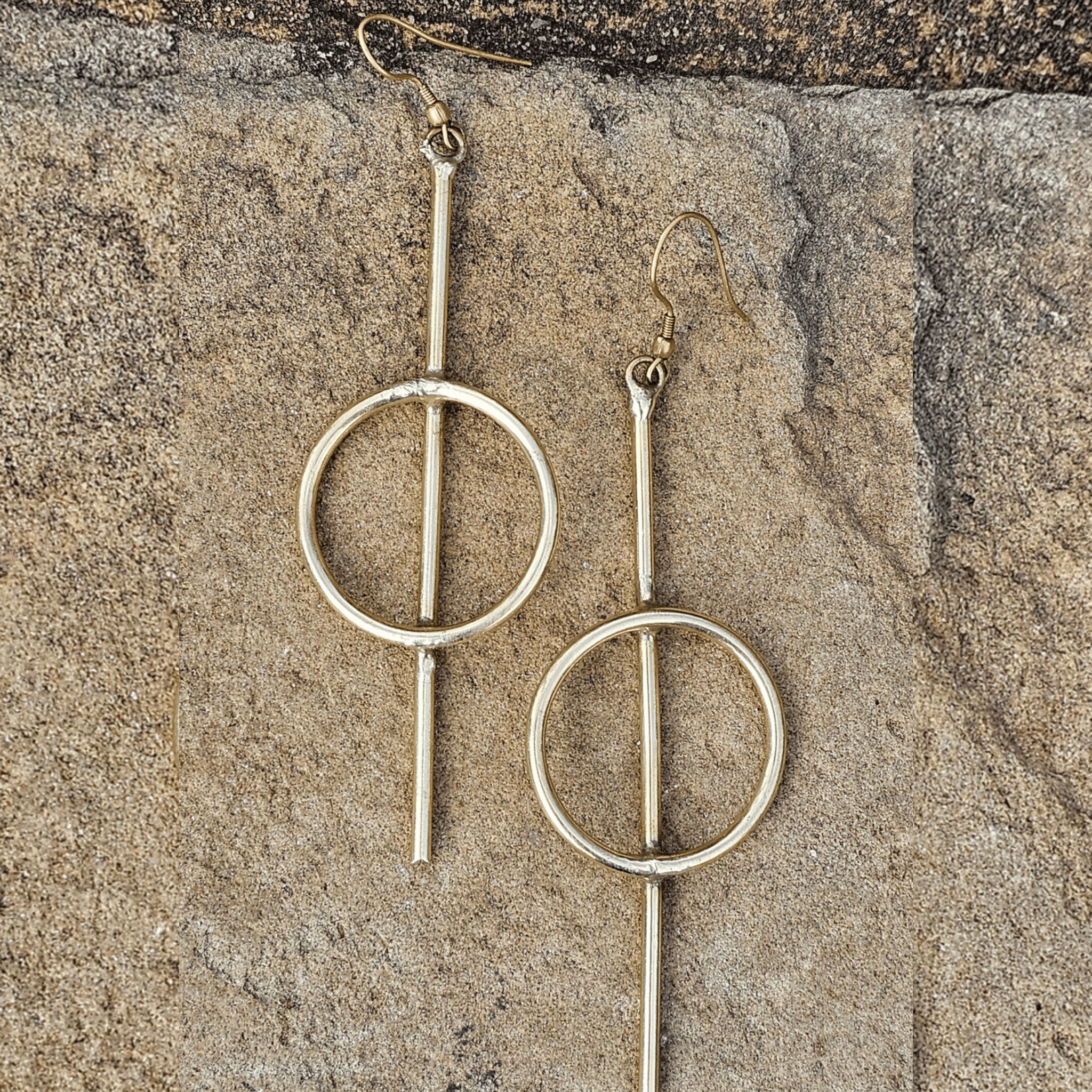 A pair of gold-toned dangle earrings with a minimalist circular design, displayed on a textured stone surface. Shop Kamawe
