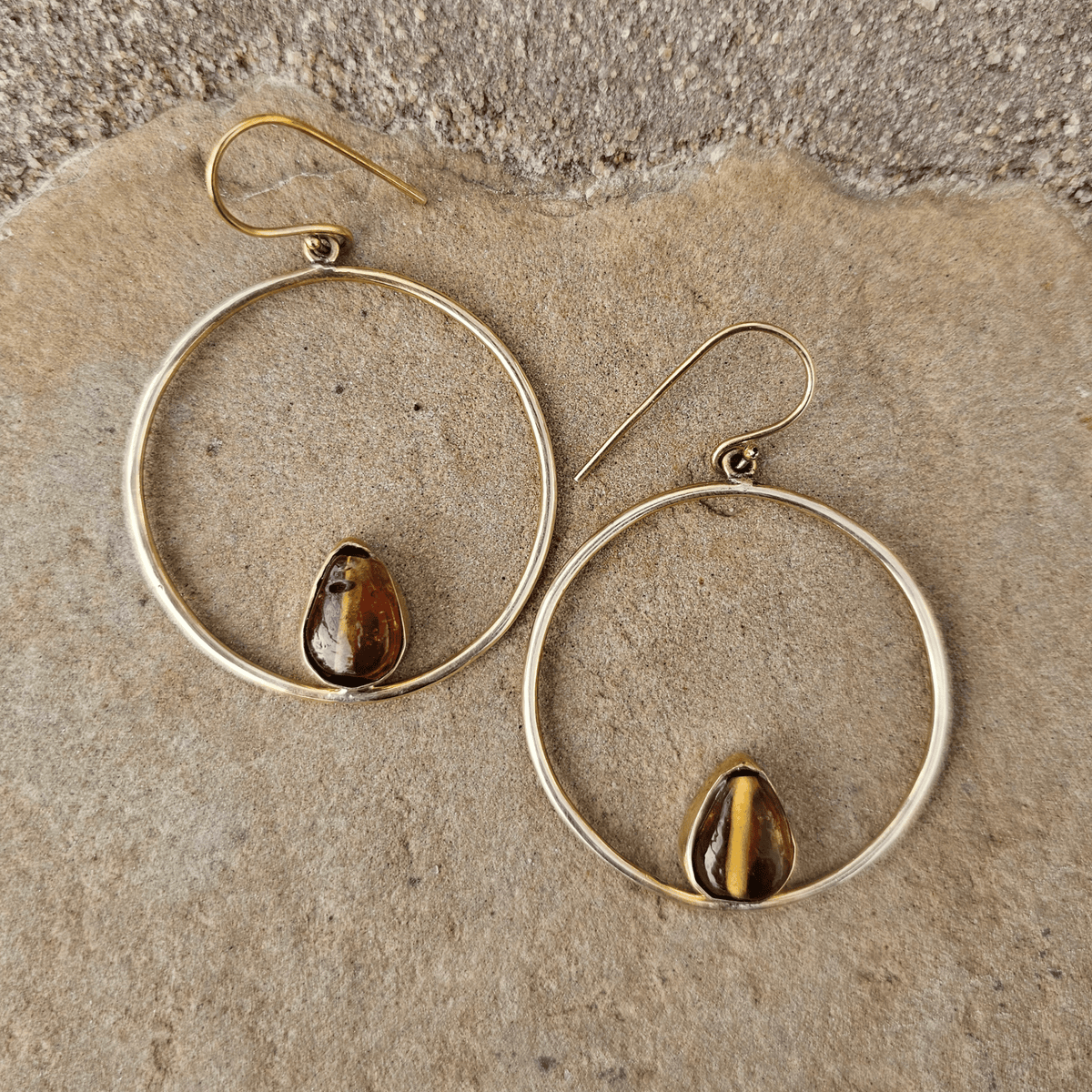A pair of gold-tone hoop earrings with small teardrop-shaped brown and gold tiger's eye stones, displayed on a textured beige stone surface. Shop Kamawe