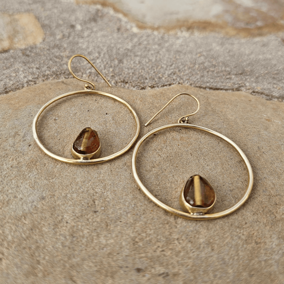 A pair of gold-toned dangle earrings with large open circular hoops and teardrop-shaped brown gemstones, displayed on a textured stone surface.