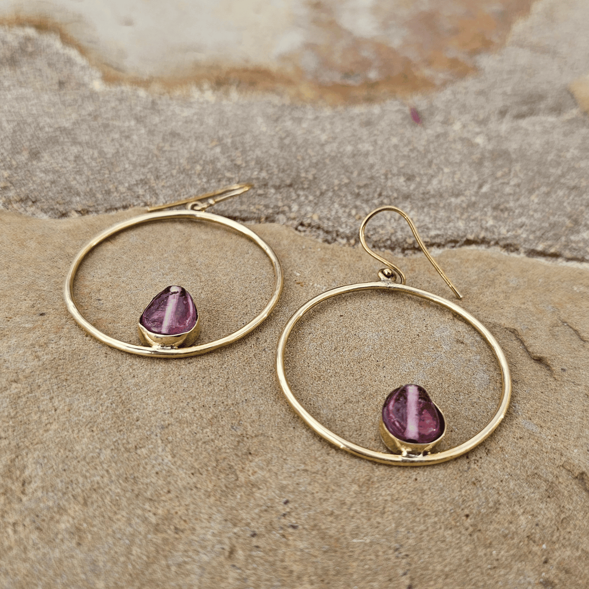 A pair of gold hoop dangle earrings with teardrop-shaped purple amethyst stones, displayed on a textured sandy surface. Shop Kamawe
