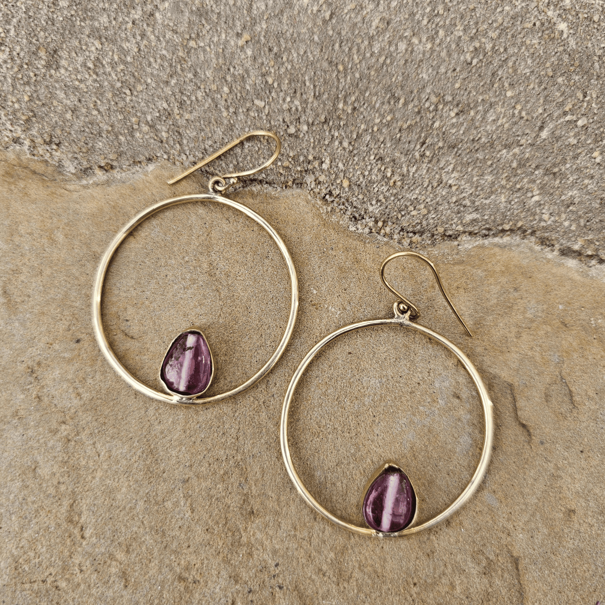 A pair of gold hoop earrings with a small teardrop-shaped purple gemstone set inside each hoop, displayed on a textured beige stone surface. Shop Kamawe