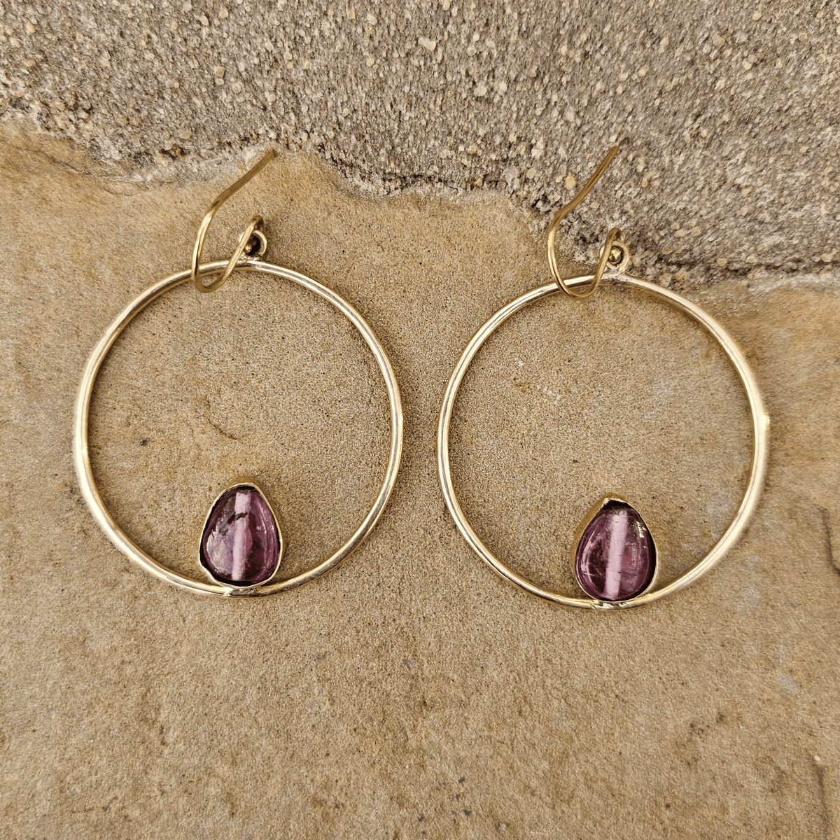 A pair of gold hoop earrings with a small teardrop-shaped purple gemstone set inside each hoop, displayed on a textured beige stone surface. Shop Kamawe