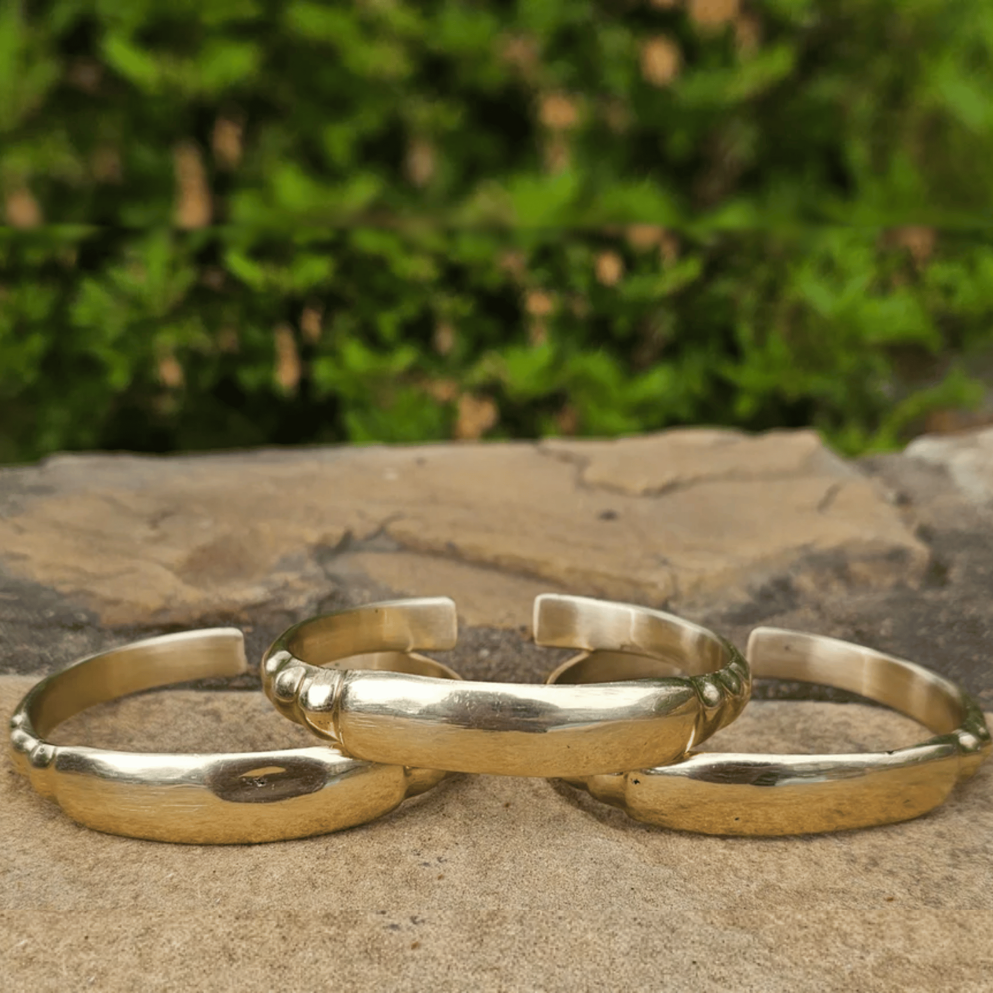 Three polished gold-toned metal bangles with textured, hammered edges are arranged on a stone surface with a blurred green outdoor background. Shop Kamawe