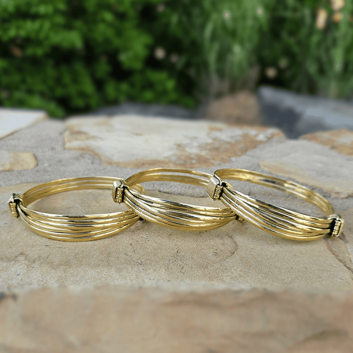 Three gold-toned textured bangles with jagged edges, displayed on a stone surface with a blurred green garden background. Shop Kamawe
