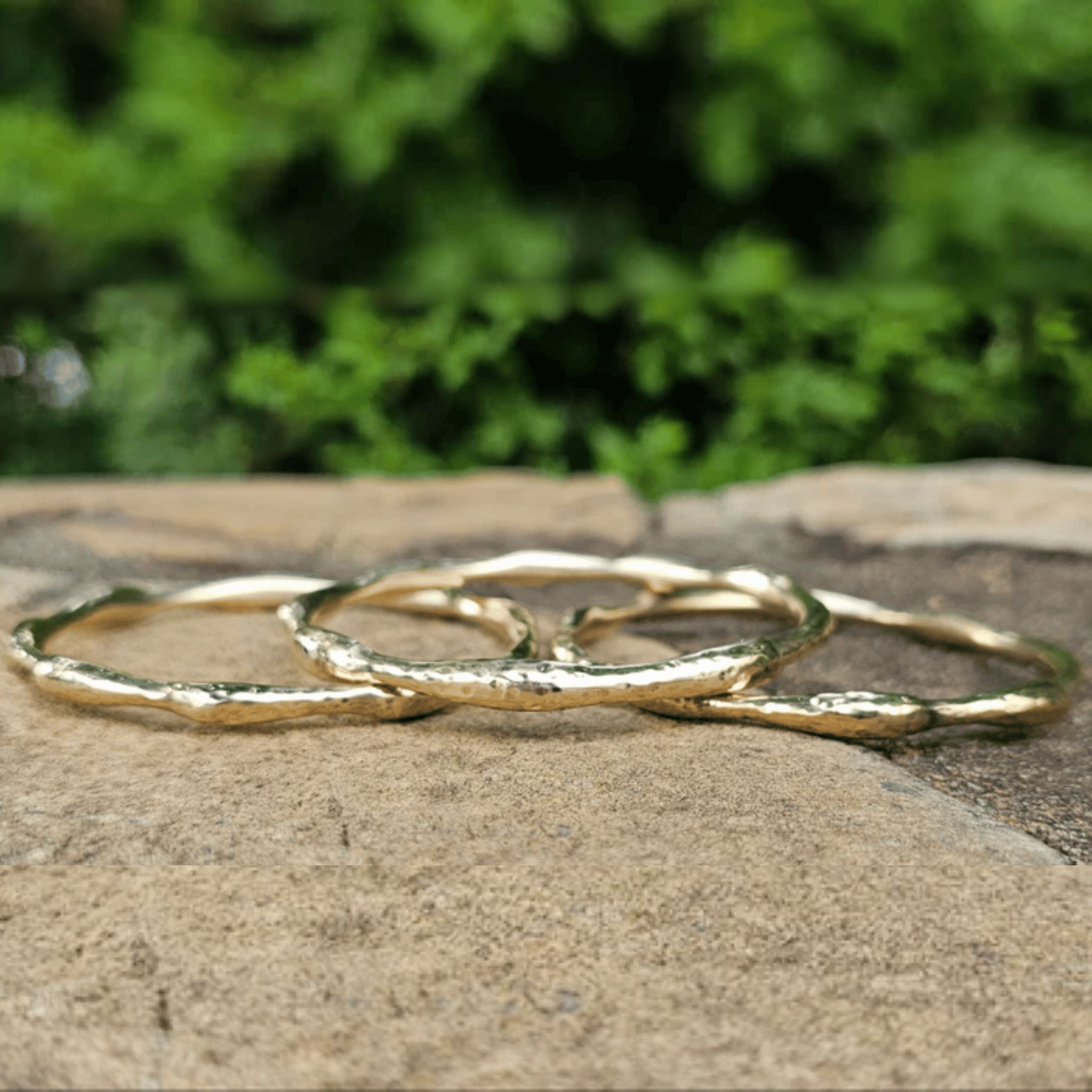 Three textured gold rings with a hammered finish, overlapping on a stone surface with a blurred green foliage background. Shop Kamawe