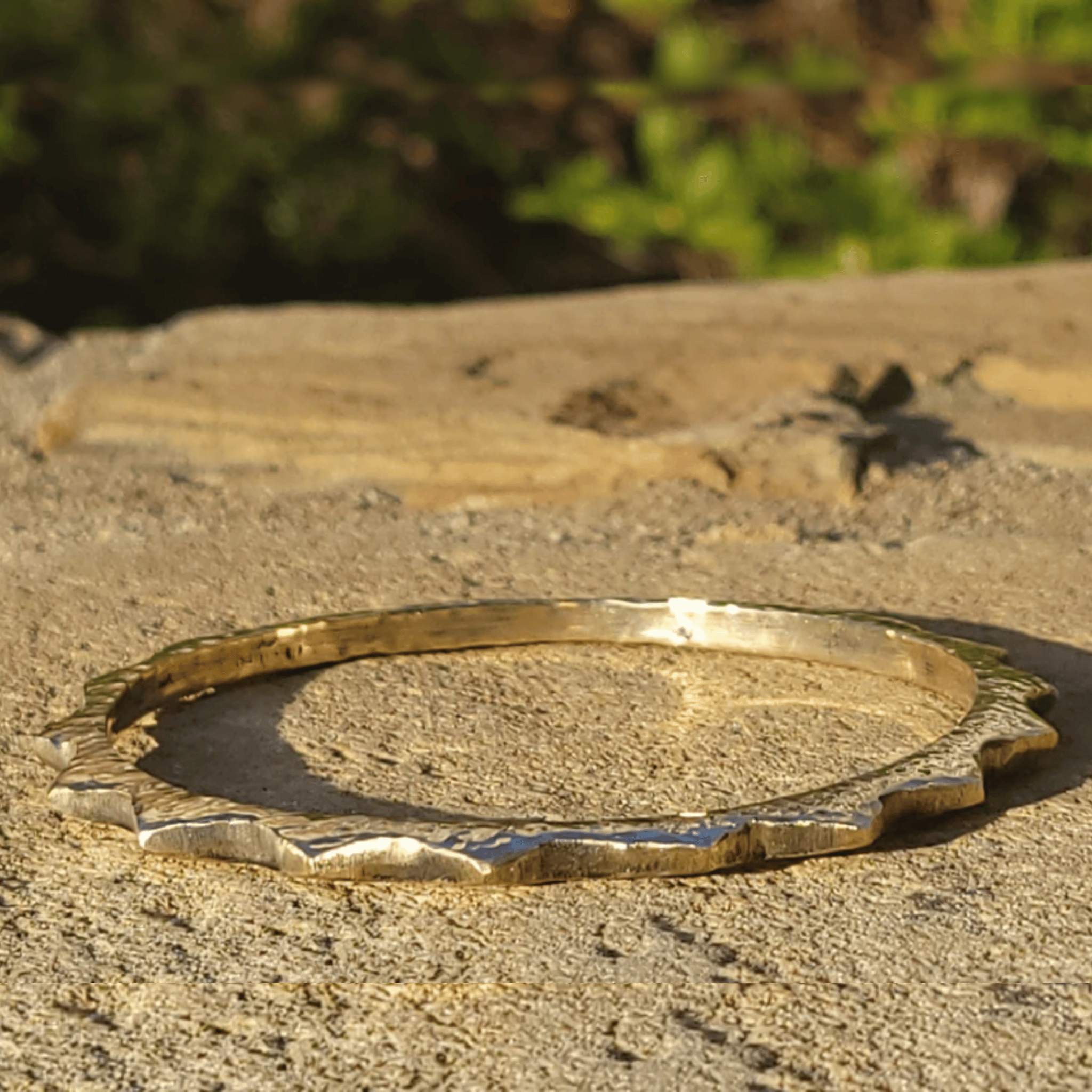 A textured gold-toned bangle bracelet with a wavy, scalloped edge, resting on a sandy surface with a blurred natural background. Shop Kamawe