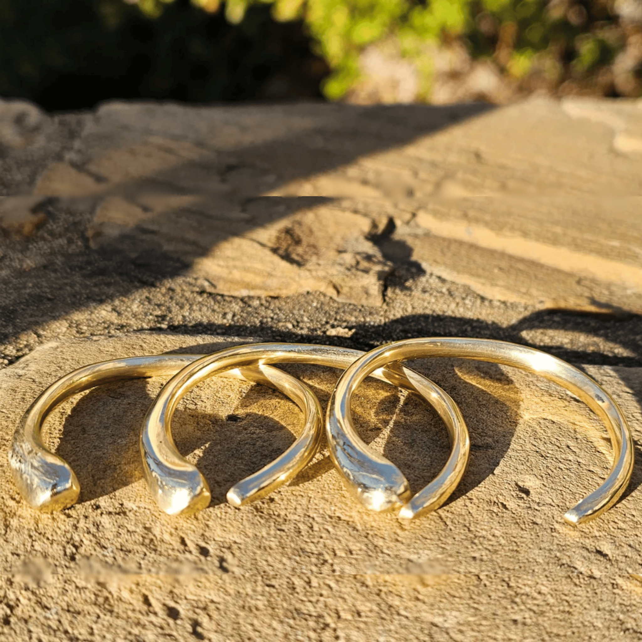 Three gold-toned curved cuff earrings arranged on a textured stone surface outdoors, with warm sunlight and blurred greenery in the background. Shop Kamawe