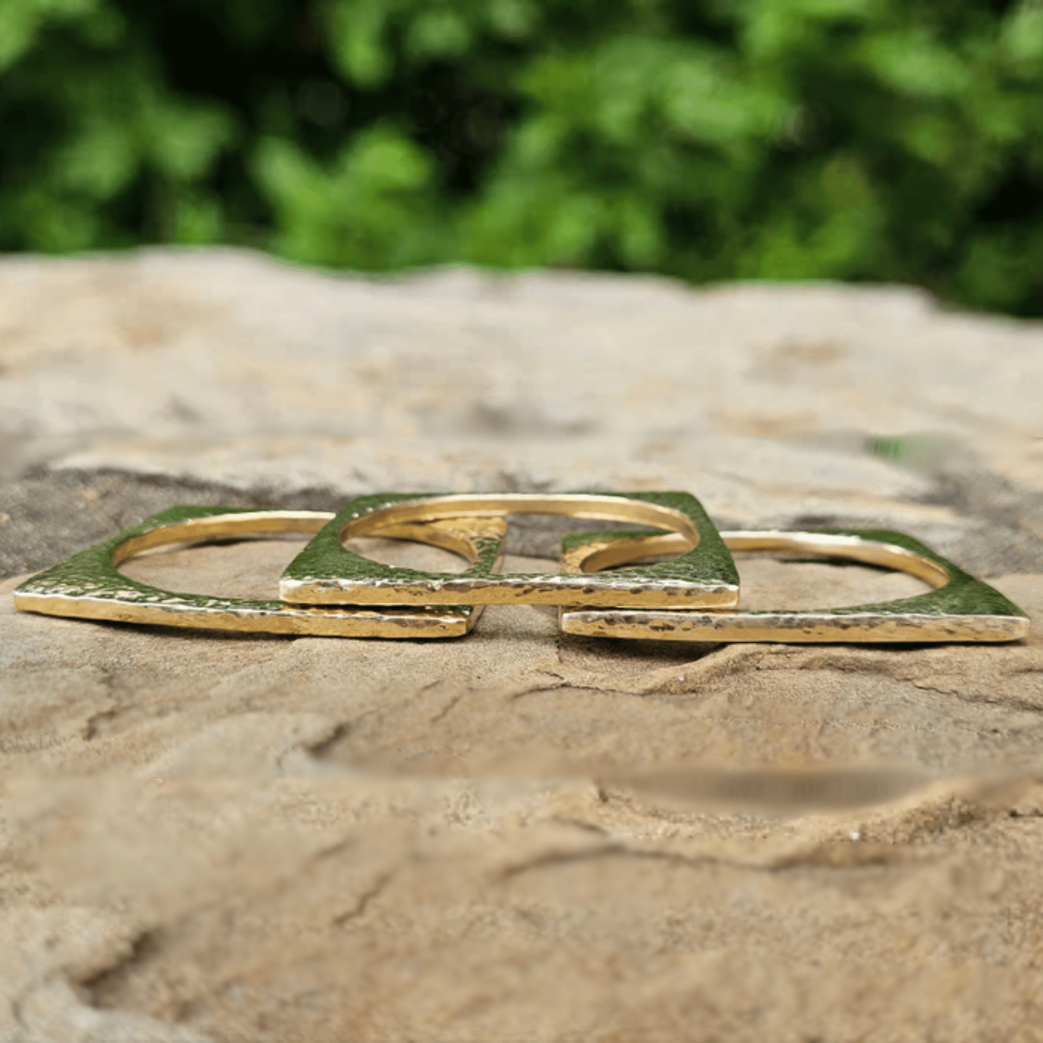 A gold-toned geometric ring with a textured finish, resting on a natural stone surface with a blurred green foliage background. Shop Kamawe