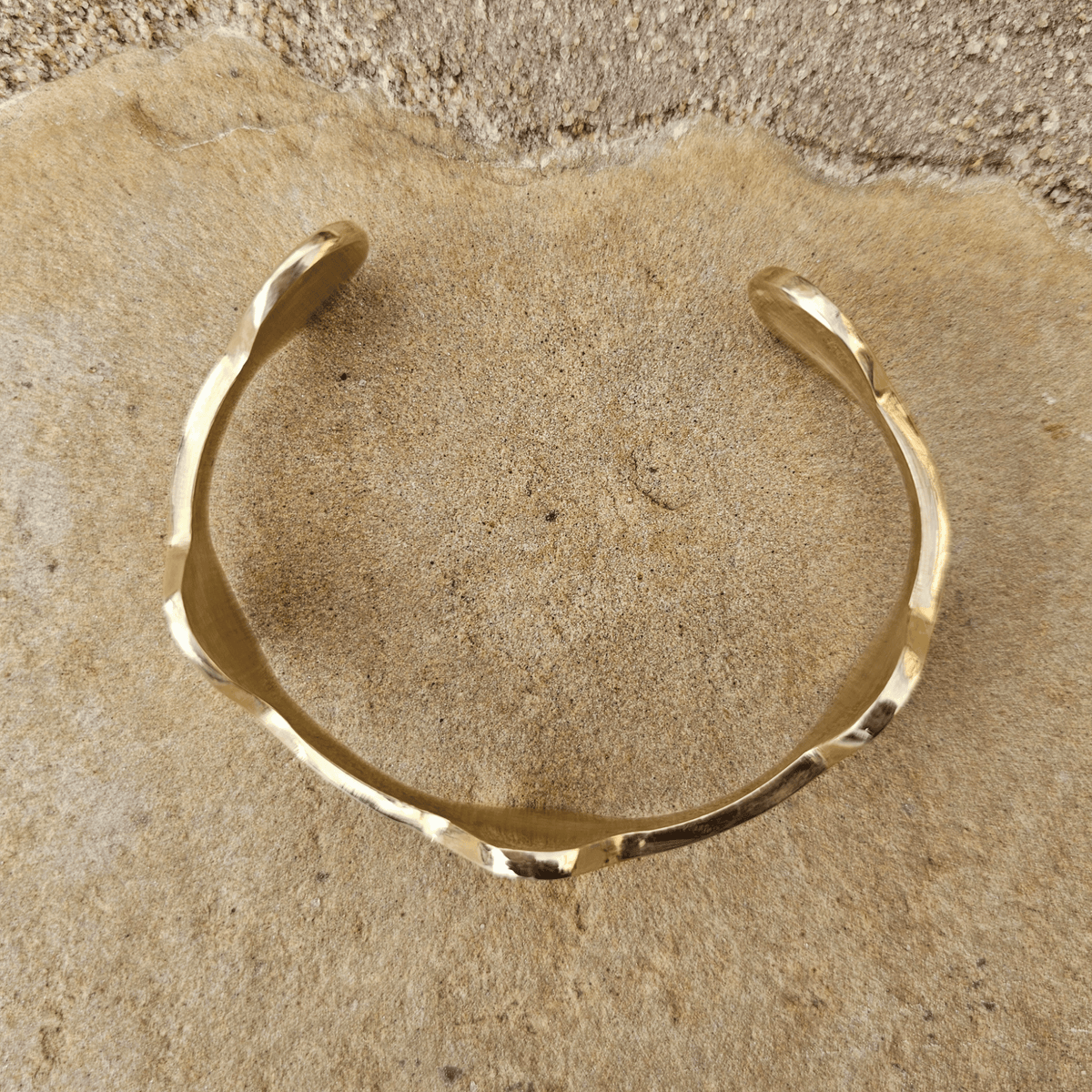 A gold-toned open cuff bracelet with organic wavy edges, resting on a textured beige stone surface. Shop Kamawe