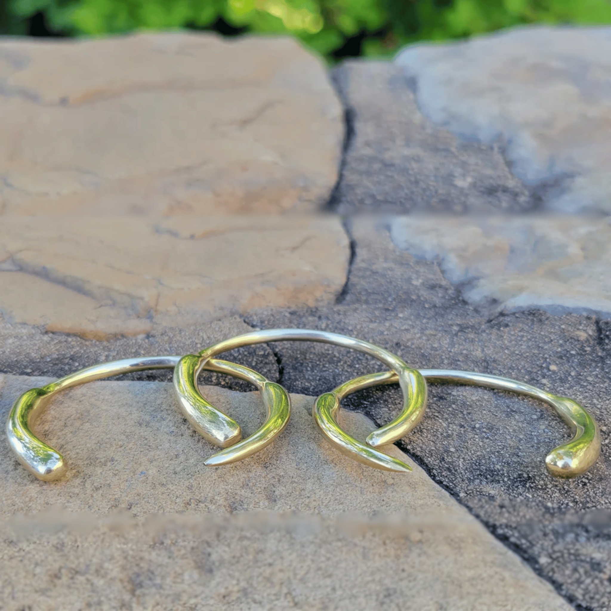 Three gold-toned wrap-around cuff earrings arranged on a stone surface outdoors. Shop Kamawe