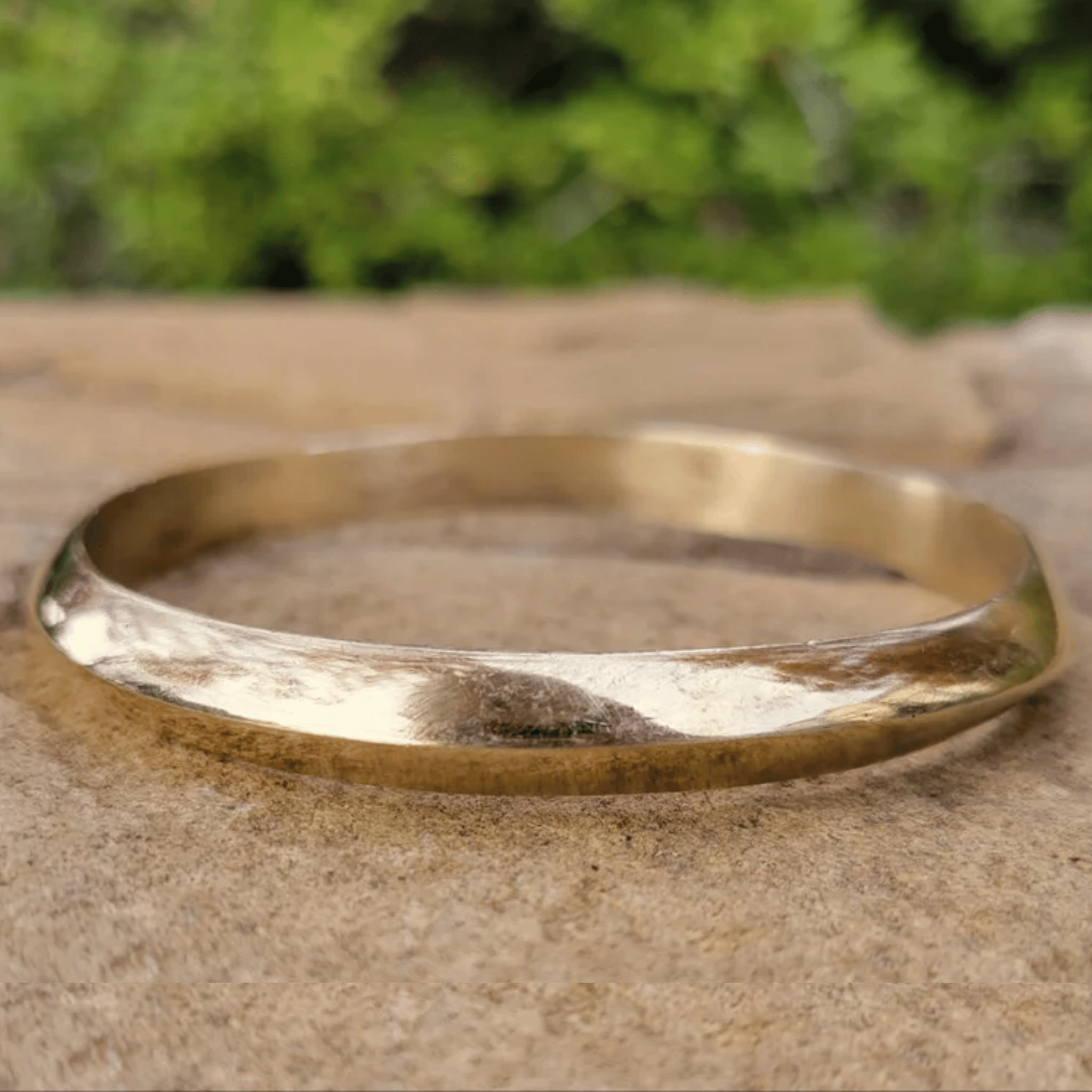 A hammered gold bangle bracelet resting on a textured stone surface with a blurred green foliage background. Shop Kamawe