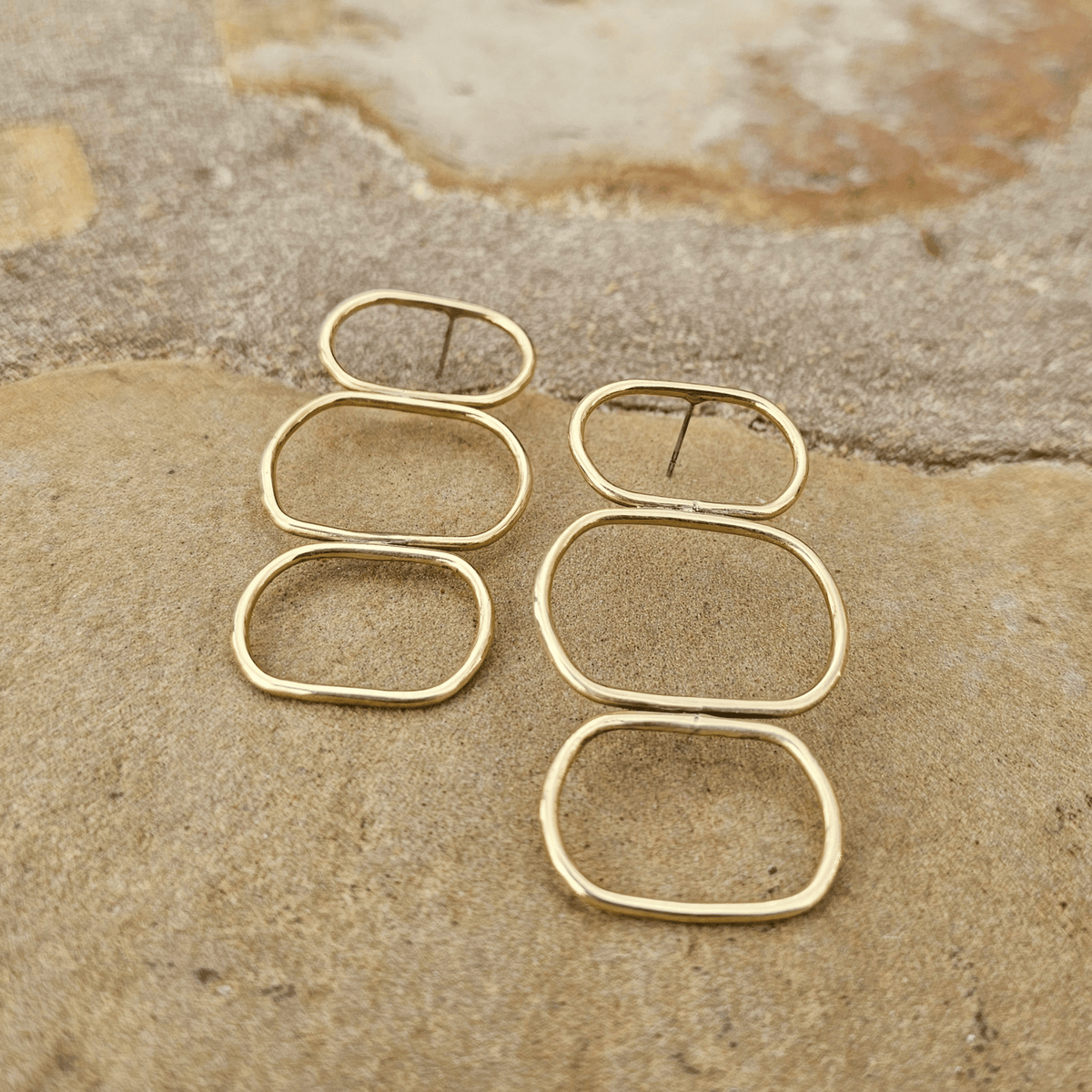 A pair of gold-toned geometric hoop earrings with elongated rounded rectangular shapes, displayed on a textured beige stone surface. Shop Kamawe