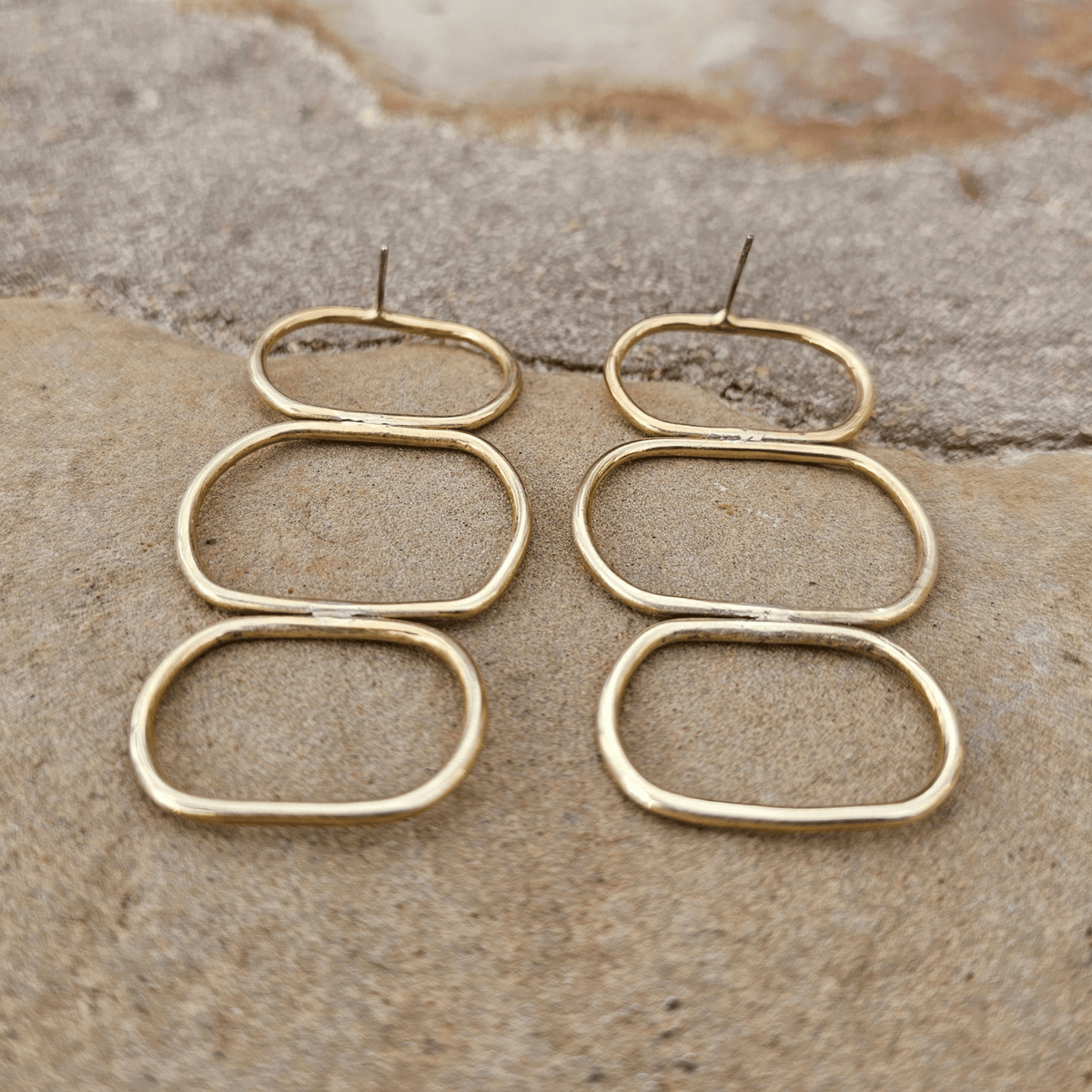 A pair of gold-toned open hoop earrings with three stacked oval rings, displayed on a textured stone surface. Shop Kamawe