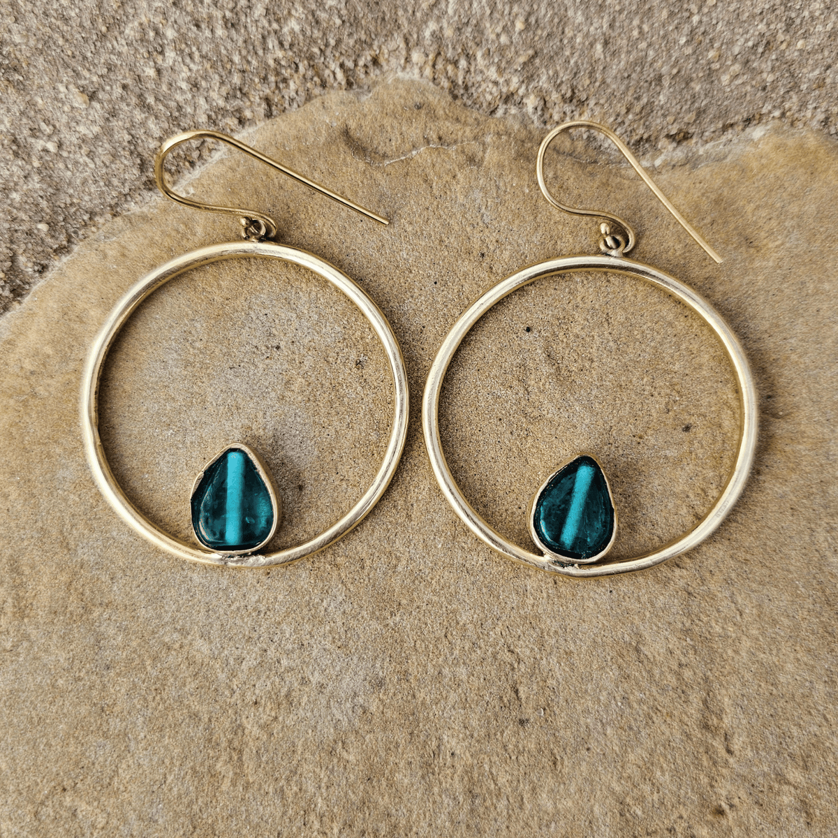 A pair of gold-tone hoop earrings with a small teardrop-shaped dark teal stone set in the lower half, displayed on a textured tan stone surface. Shop Kamawe