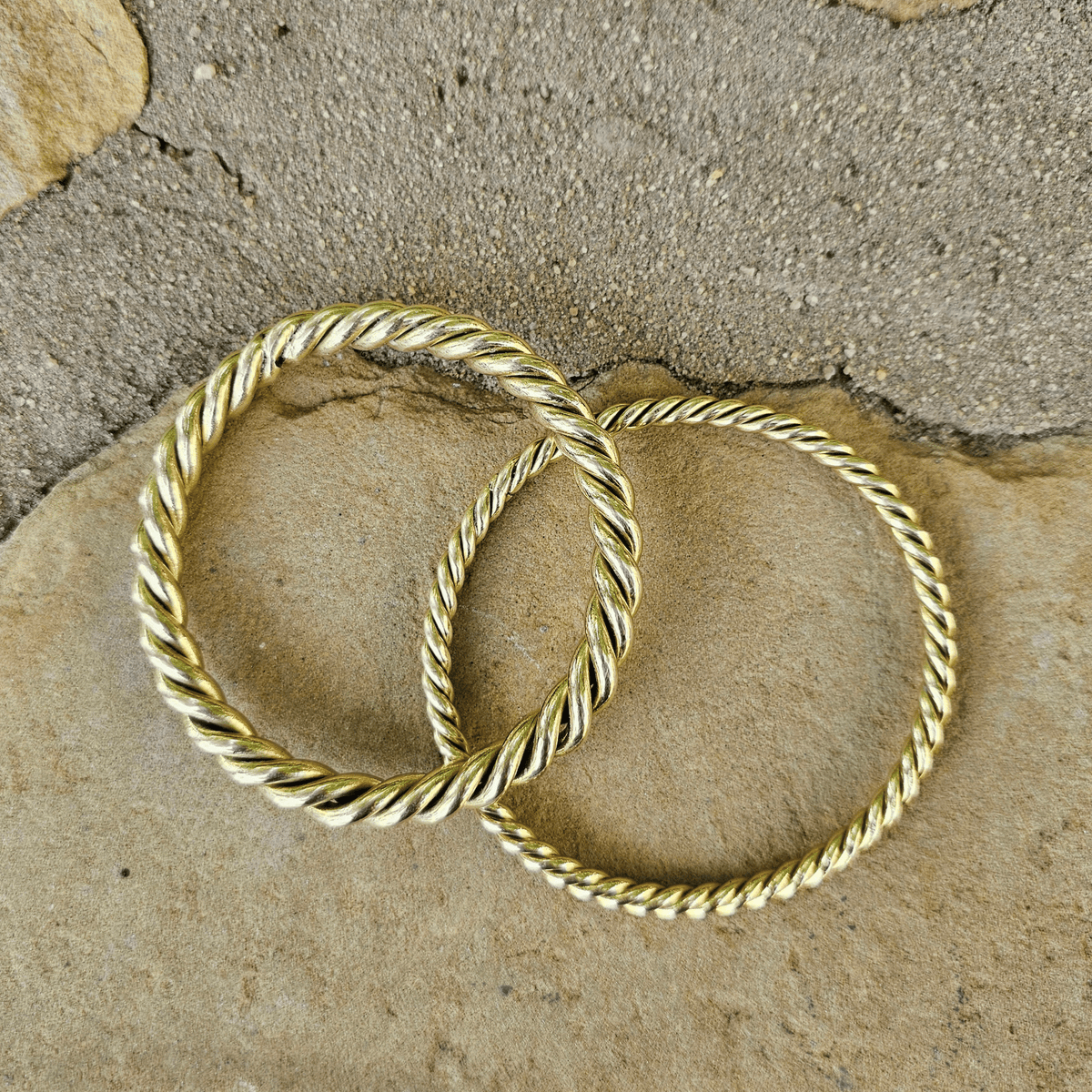 Two gold-toned twisted rope bangles resting on natural tan and gray stones. Shop Kamawe