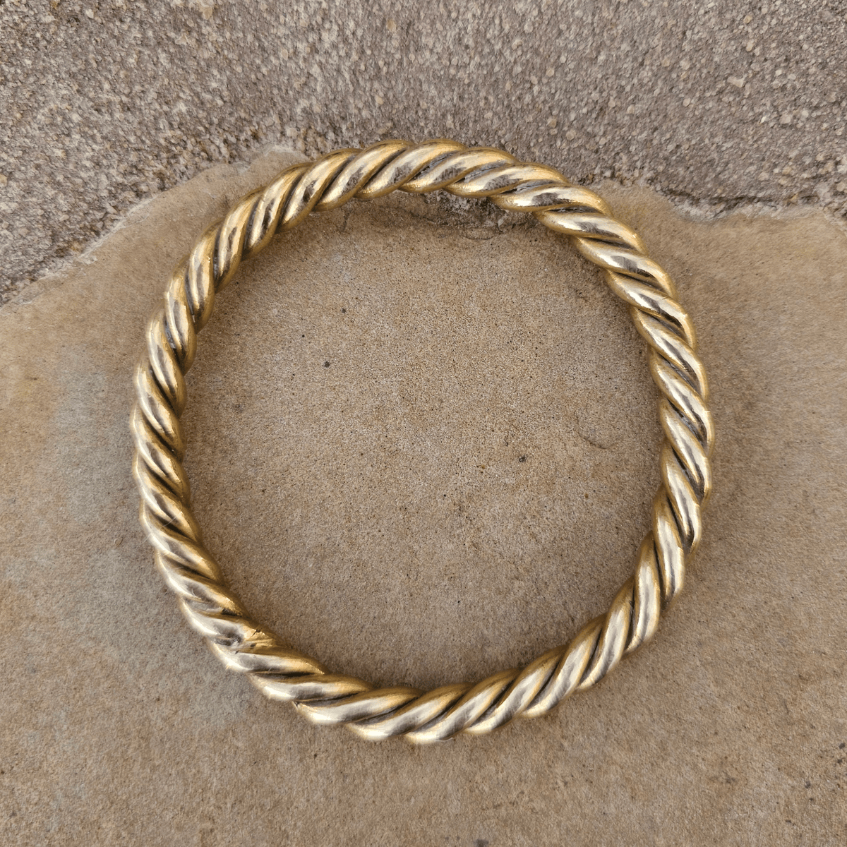 A twisted rope-style gold bangle bracelet resting on a textured tan stone surface. Shop Kamawe
