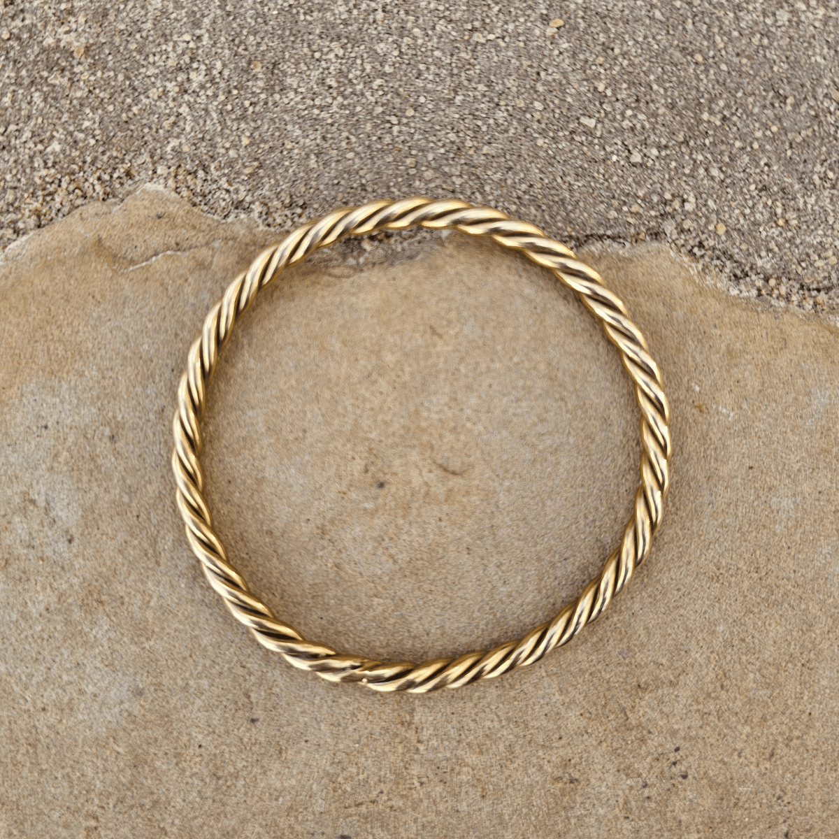 A gold-toned twisted rope bangle bracelet resting on a textured stone surface. Shop Kamawe