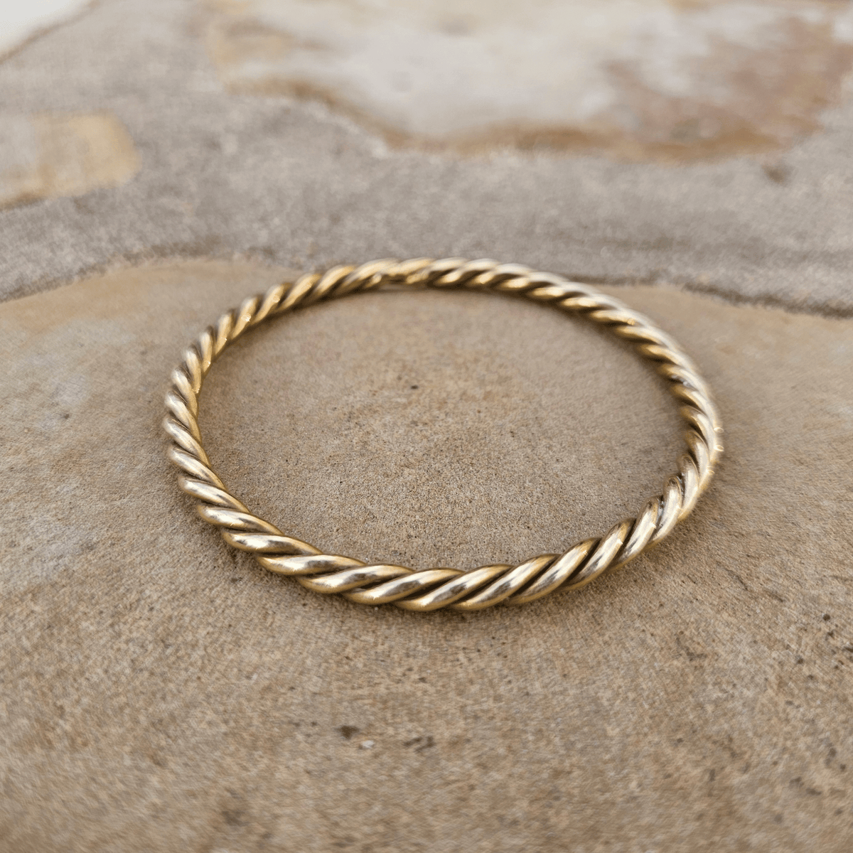 A thin twisted rope-style gold bangle bracelet resting on a textured beige stone surface. Shop Kamawe