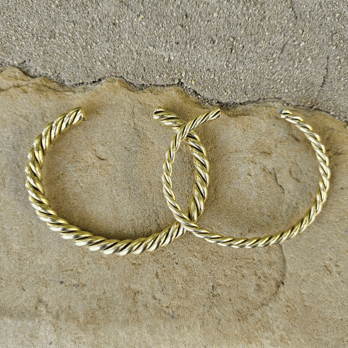 A pair of gold-toned twisted hoop earrings resting on a textured beige stone surface. Shop Kamawe
