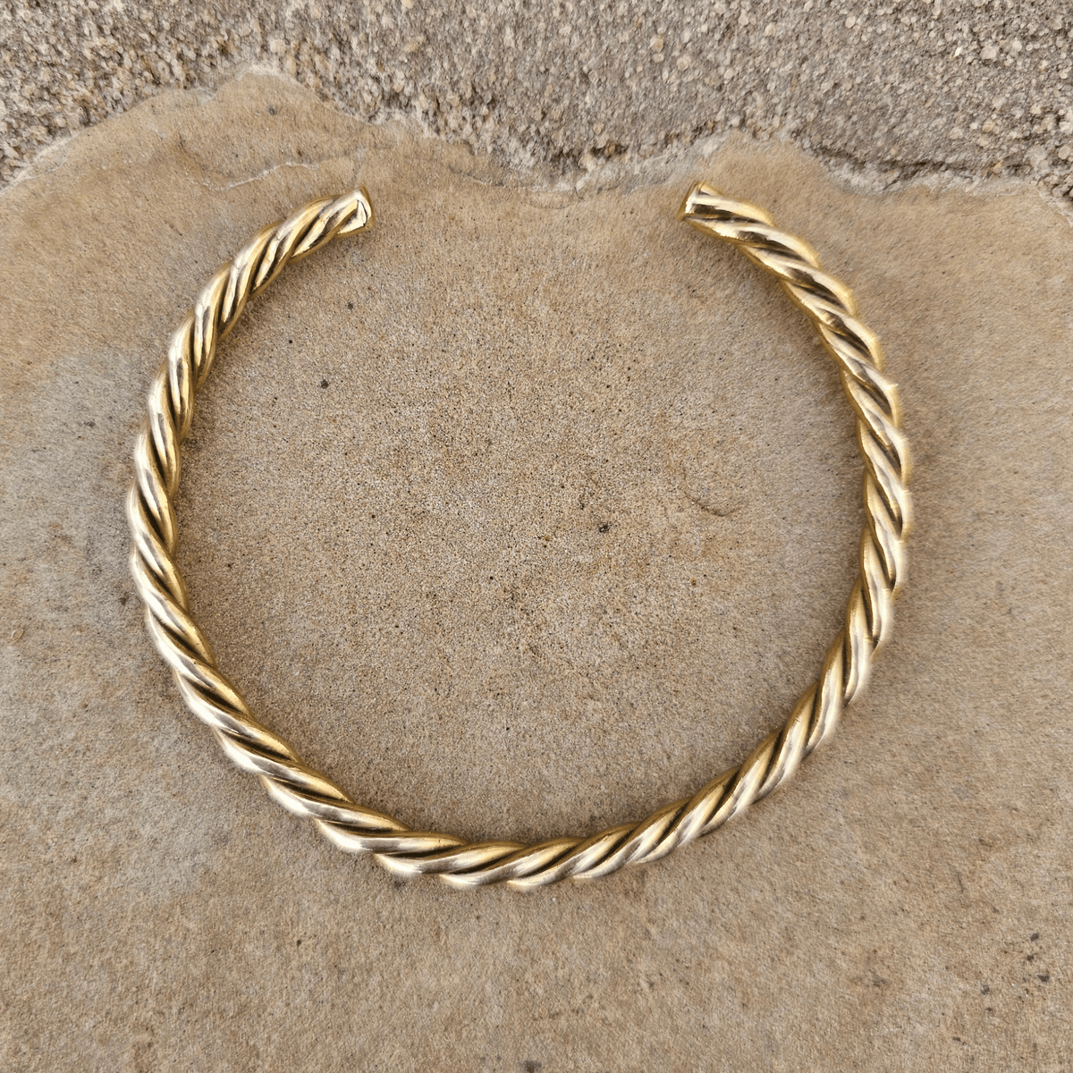 A twisted gold-tone metal cuff bracelet, likely made of copper or gold, displayed on a textured tan stone surface. Shop Kamawe