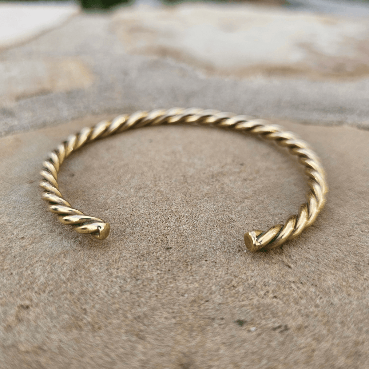 A twisted rope-style open cuff bracelet in polished yellow gold, resting on a textured stone surface. Shop Kamawe