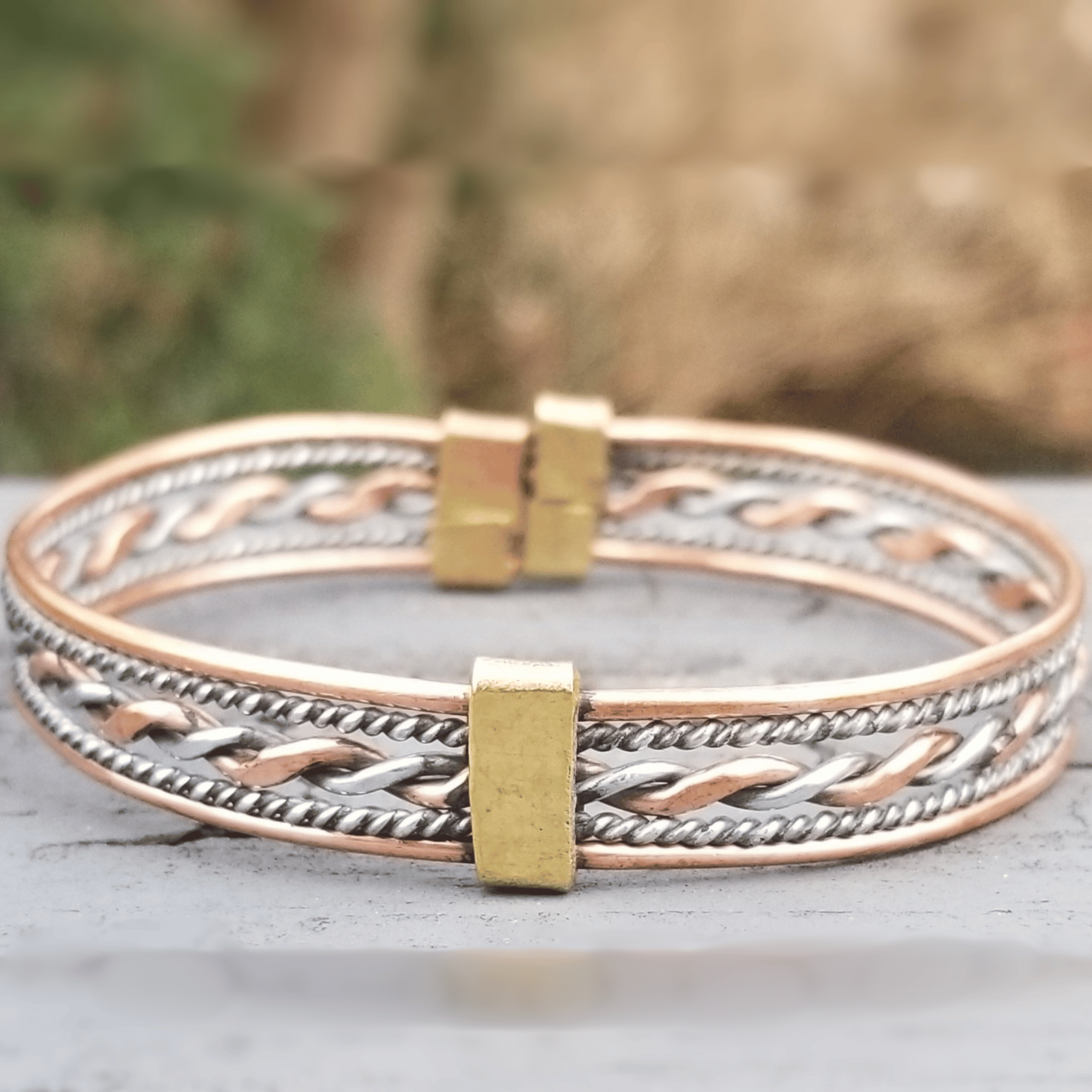 A close-up of a multi-strand bangle bracelet featuring twisted copper and silver rope patterns with a central gold-toned rectangular accent, resting on a gray surface with a blurred natural background. Shop Kamawe