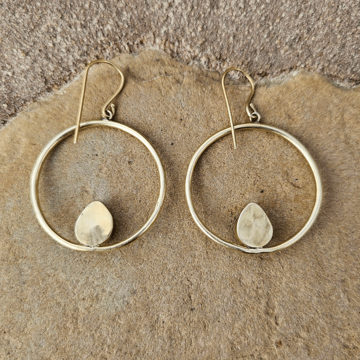 A pair of gold-tone hoop earrings with a small teardrop-shaped yellow-green bead or stone set inside each hoop, displayed on a textured beige stone surface. Shop Kamawe