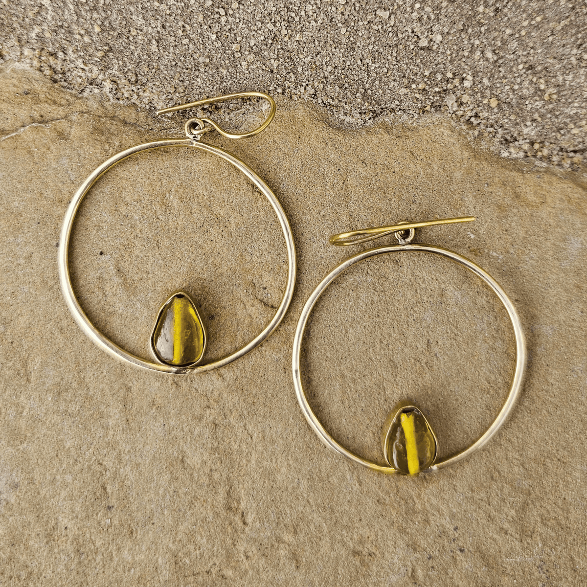 A pair of gold-tone hoop earrings with a small teardrop-shaped yellow-green bead or stone set inside each hoop, displayed on a textured beige stone surface. Shop Kamawe