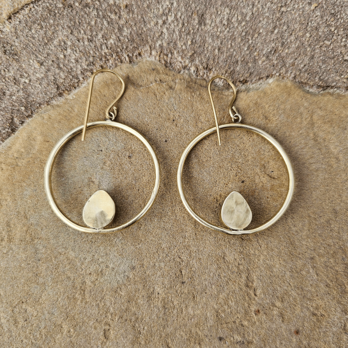 A pair of gold-tone hoop earrings with a small teardrop-shaped stone set in the center of each hoop, displayed on a textured natural stone surface. Shop Kamawe