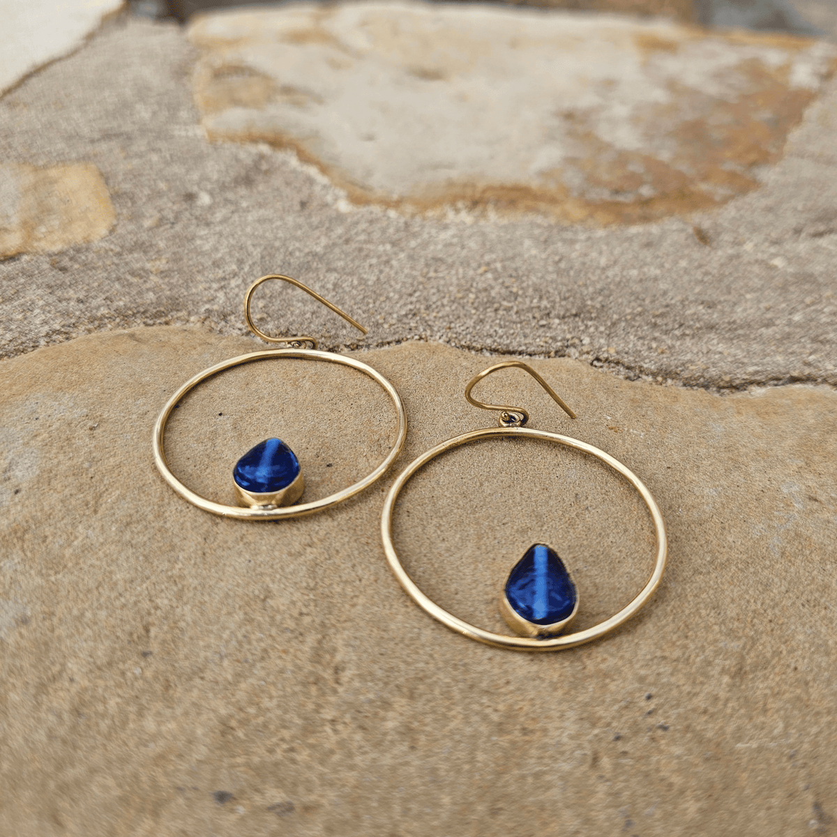 A pair of gold hoop dangle earrings with teardrop-shaped blue gemstones, displayed on a textured beige stone surface. Shop Kamawe