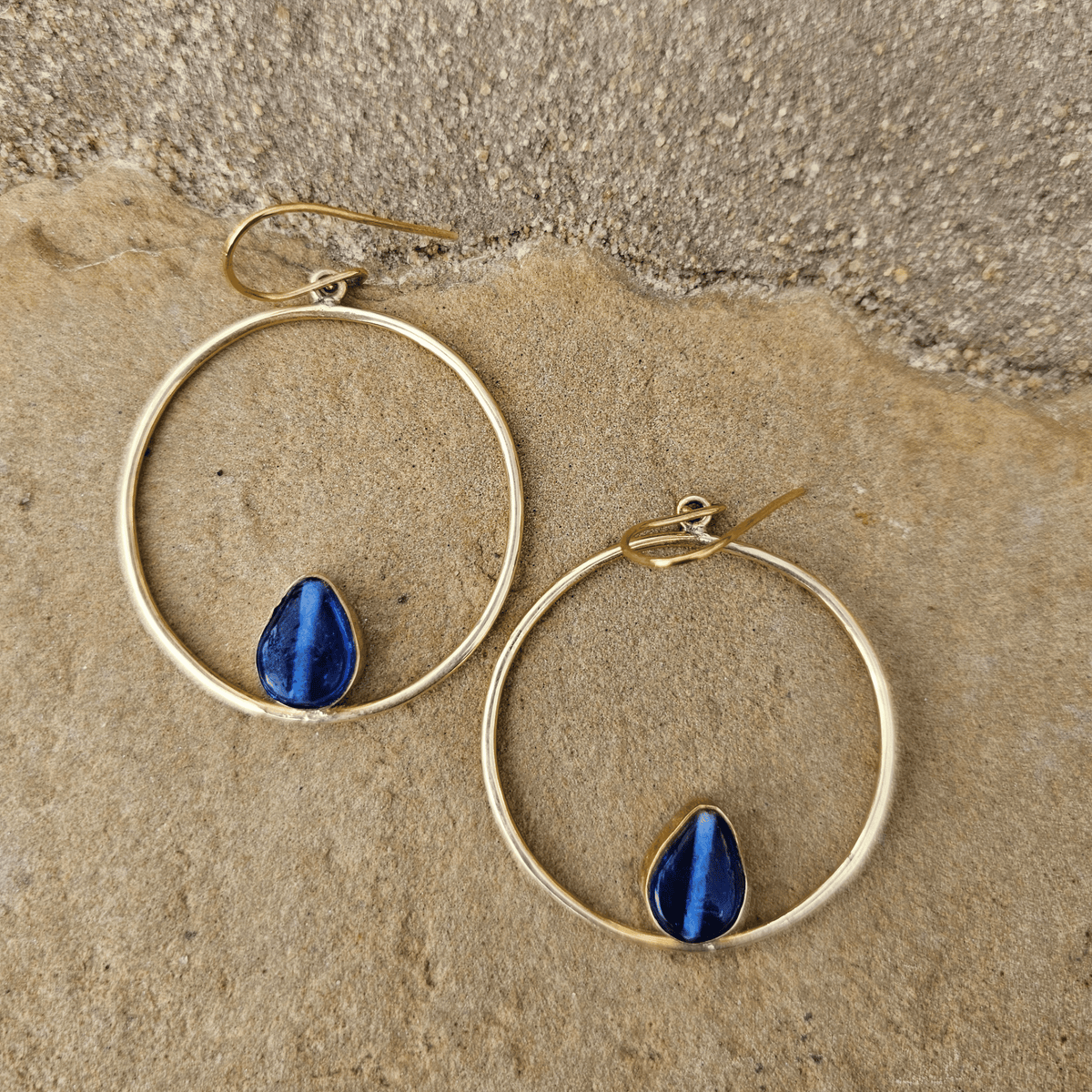 Gold hoop earrings with teardrop-shaped blue lapis lazuli stones, displayed on a textured beige stone surface. Shop Kamawe