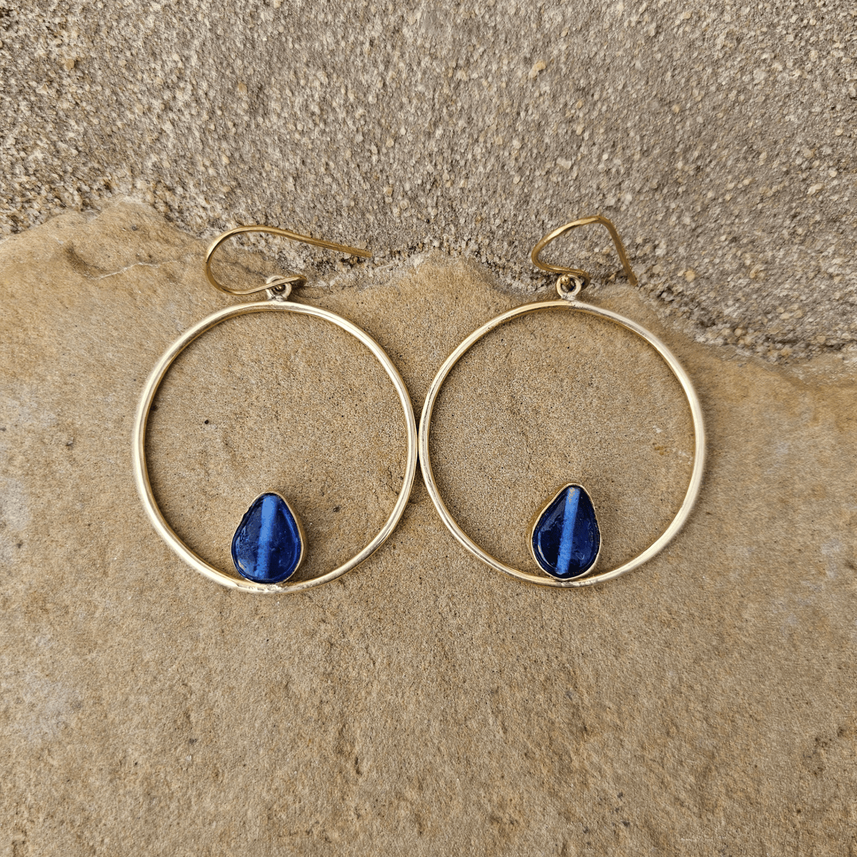 Gold hoop earrings with teardrop-shaped blue lapis lazuli stones, displayed on a textured beige stone surface. Shop Kamawe