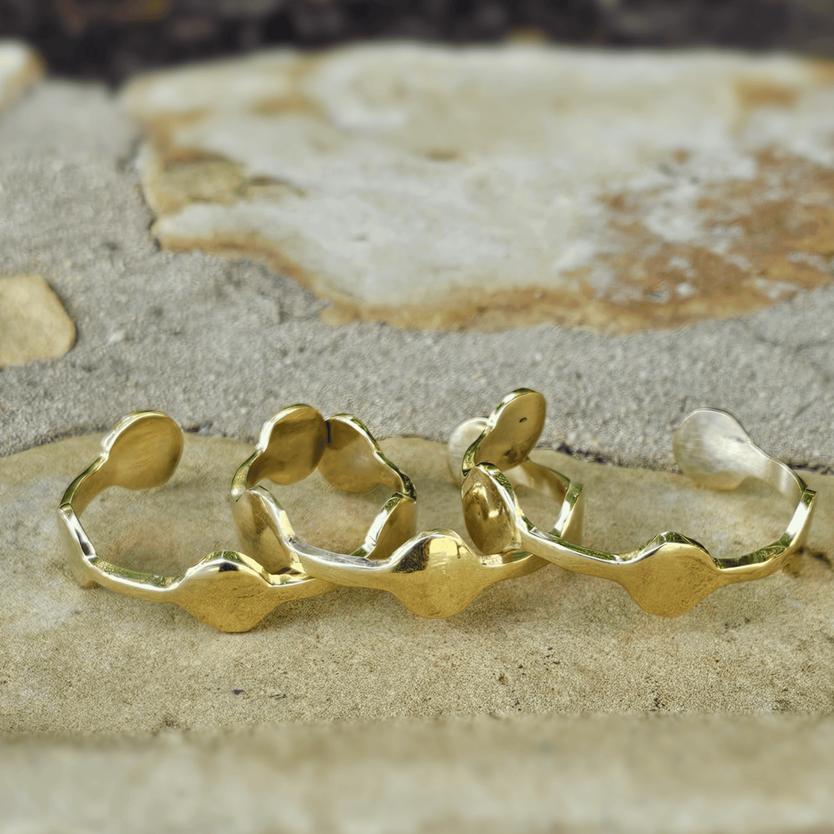 Three gold-toned wavy open cuff rings arranged on a textured sandy surface with a blurred natural stone background. Shop Kamawe