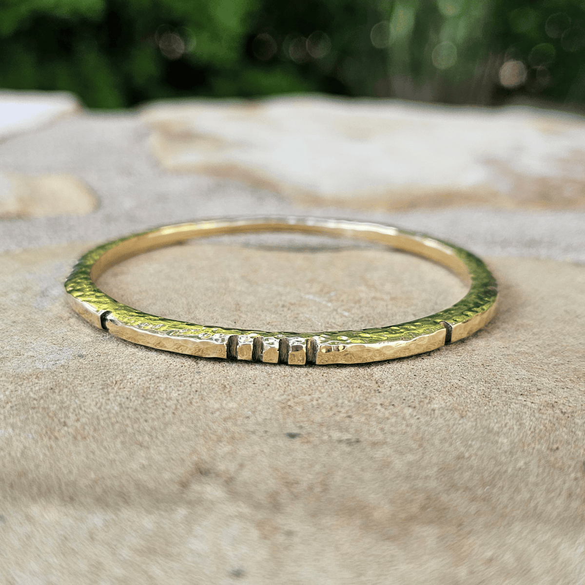 A thin gold bangle bracelet with a textured hammered finish and three small raised segments on one side, resting on a beige stone surface with a blurred green background. Shop Kamawe