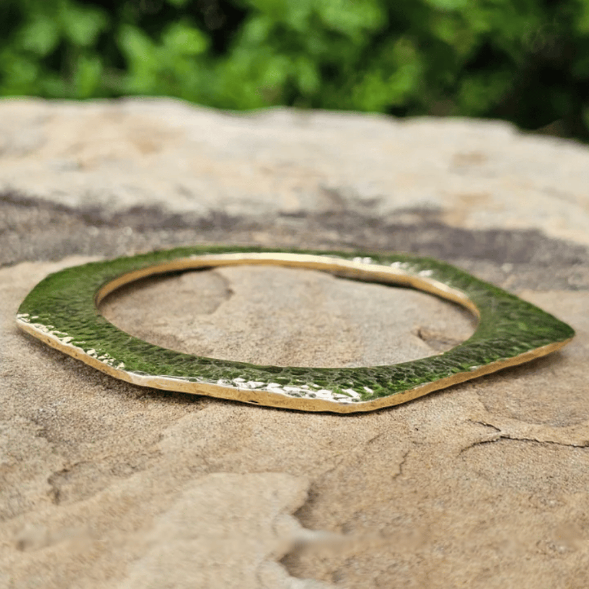 A hammered green and gold metal cuff bracelet resting on a textured stone surface with a blurred green garden background. Shop Kamawe