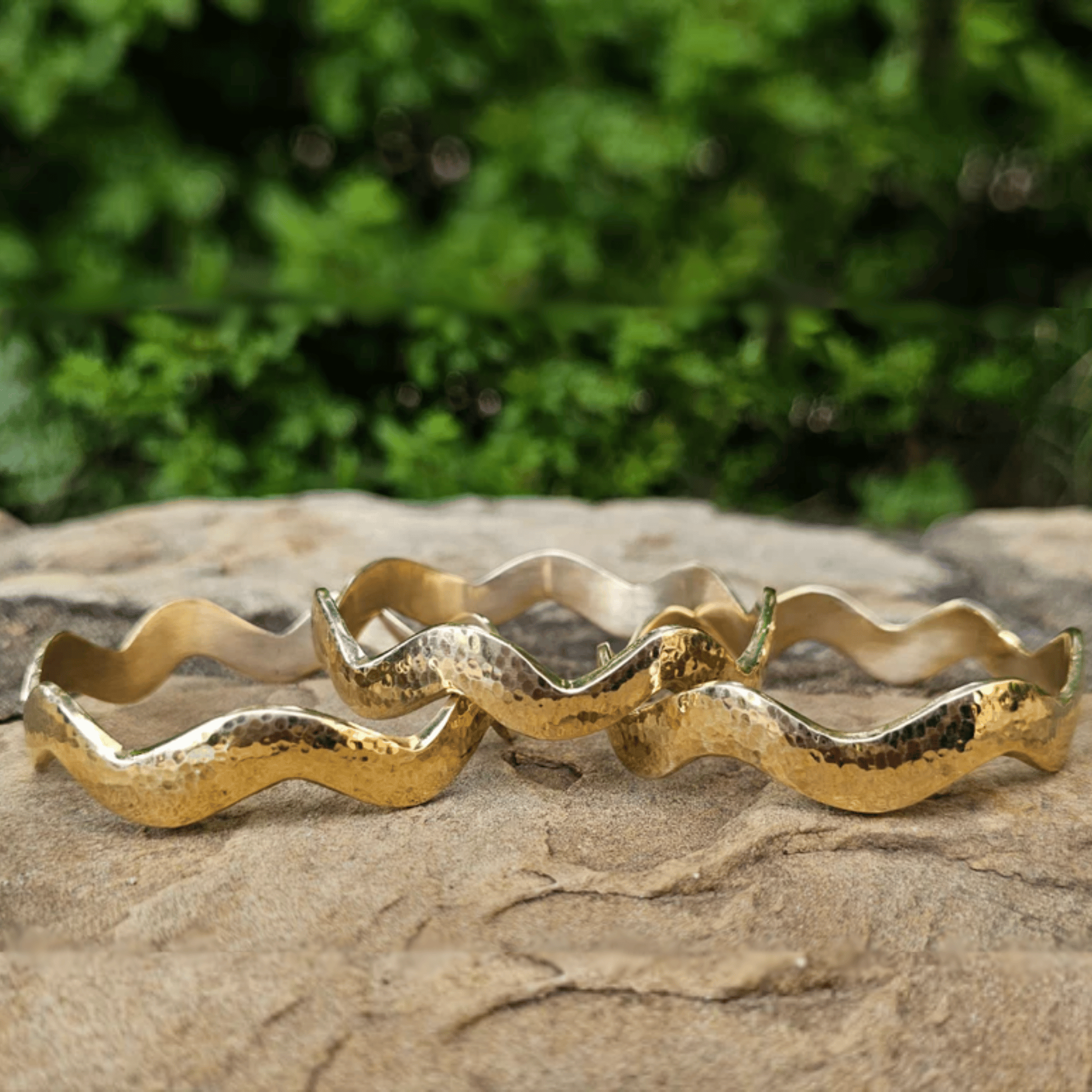 Three hammered gold wavy bangles displayed on a textured stone surface with a blurred green foliage background. Shop Kamawe