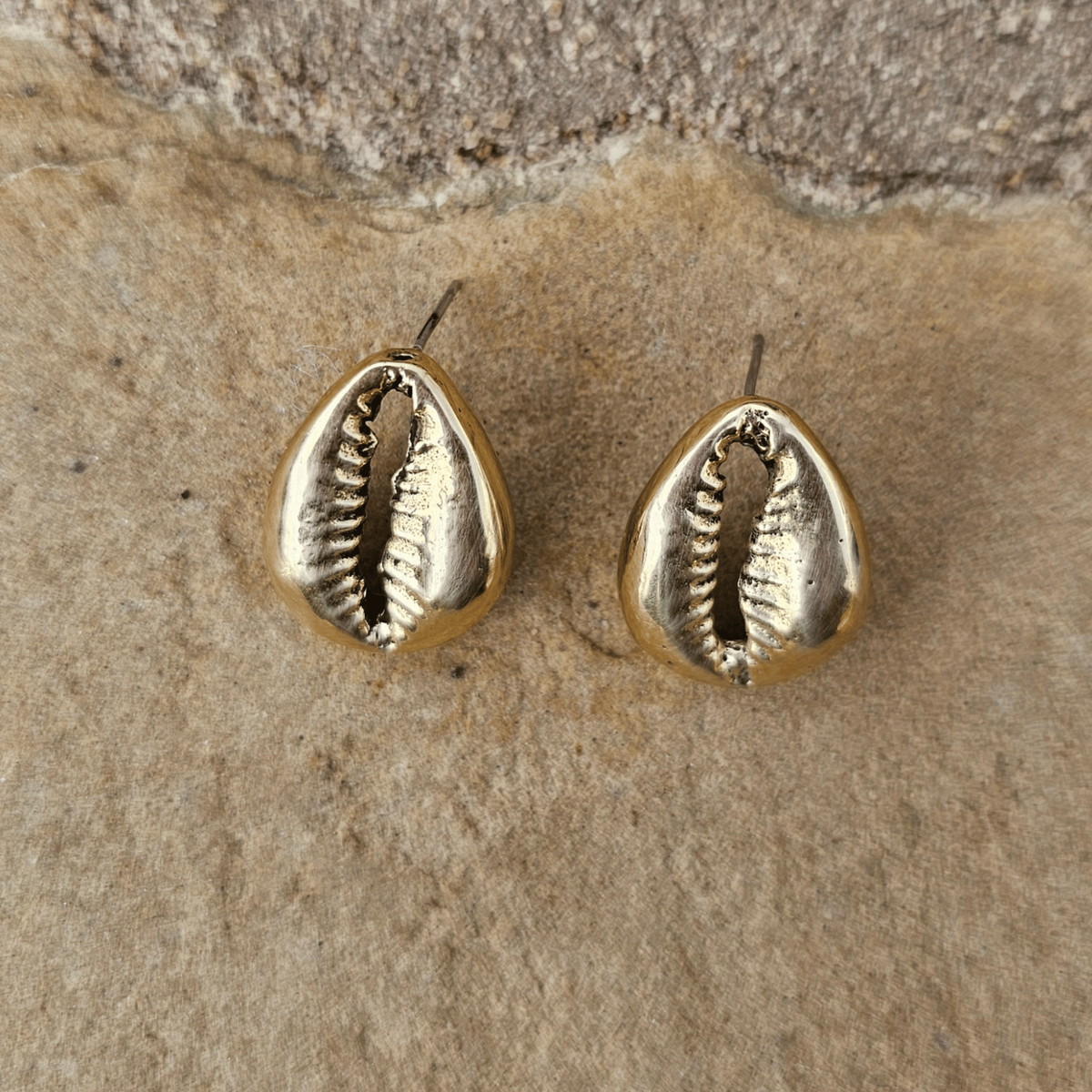 A pair of gold-toned cowrie shell stud earrings resting on a textured beige stone surface. Shop Kamawe