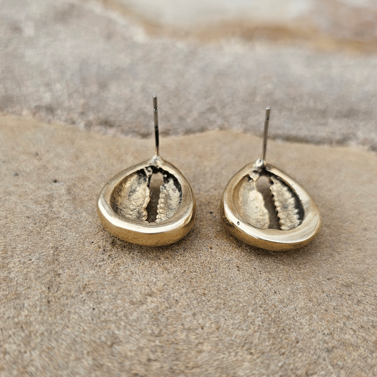 A pair of small gold-toned stud earrings with a hammered metallic finish, displayed on a textured stone surface. Shop Kamawe