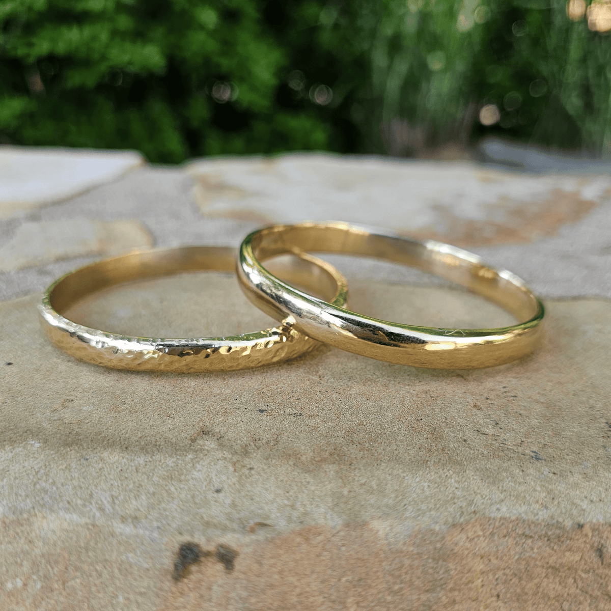 Two gold bands, one with a hammered texture and one smooth, resting on a stone surface with a blurred green garden background. Shop Kamawe